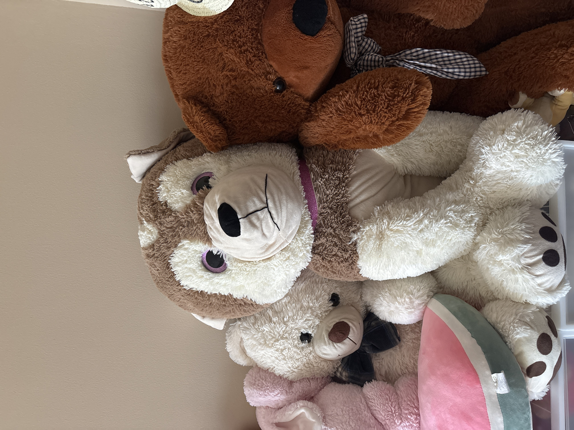 A collection of plush toys are arranged against a light beige wall. From left to right, a pink plush bunny toy is partially visible. Next to it is a large, fluffy white teddy bear wearing a dark plaid bow tie. In the center is a large stuffed husky dog with brown and white fur, large purple eyes, and a pink collar. To the right of the husky is a large brown teddy bear wearing a similar dark plaid bow tie. In the foreground, a watermelon slice-shaped pillow rests on top of the white teddy bear's paws. The toys are positioned closely together, indicating a shared space. The overall scene is domestic and appears to be a close-up shot of a shelf or surface holding these stuffed animals. There is no identifiable information to determine a specific location within Dubai, United Arab Emirates.