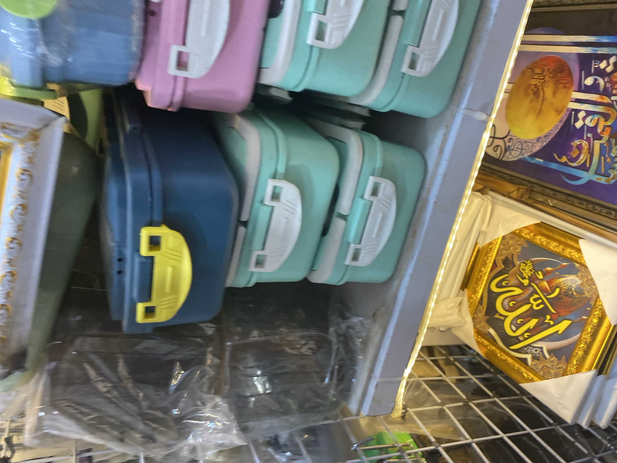 The image shows a retail shelf display of various items, primarily food containers and framed artwork. In the foreground, a framed piece of artwork with golden embellishments and Arabic calligraphy is visible. Next to it, another framed artwork with blue and purple hues and similar calligraphy is partially obscured.
Above the artwork, several stackable food containers are arranged on a shelf. There are blue containers with yellow latches, pink containers with white latches, and several light teal-colored containers, also with white latches, stacked in different configurations. Some of the containers appear to be made of plastic.
Behind the containers, a clear plastic-wrapped dark item, possibly another product or packaging, is visible on the left side of the shelf. The shelf itself is illuminated by a string of small LED lights running along its front edge. The background suggests a retail or storage environment with additional shelves and possibly other products. The overall impression is of a product display in a shop. The location is specified as Lamteumen Barat, Indonesia, suggesting this is likely a scene from a marketplace or store in that area. There are no people or actions depicted in the image.