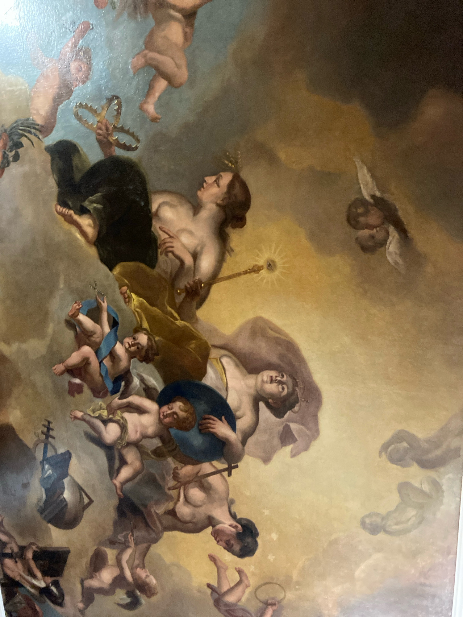 The image depicts a ceiling fresco, likely a religious or allegorical scene, featuring multiple cherubic figures and adult figures amidst clouds. The style suggests Baroque or Neoclassical influences. One prominent adult female figure is shown reclining, holding a scepter with an eye-like symbol at its tip. Another adult female figure is partially visible, looking upwards. Several cherubic figures are seen in various poses, some interacting with each other, others holding objects such as a crown-like wreath and a blue ribbon. Various objects are scattered throughout the scene, including what appears to be a ship's mast and rigging on the left side, and musical instruments. The overall color palette is warm, dominated by golden hues, browns, and pale blues, creating a dreamlike or celestial atmosphere. The painting's detailed rendering and dynamic composition suggest it is part of a larger decorative scheme in a grand building.
While I can describe the painting in detail, I cannot pinpoint its exact location in Saint Petersburg, Russia, or confirm it is indeed from that city without additional information or context.