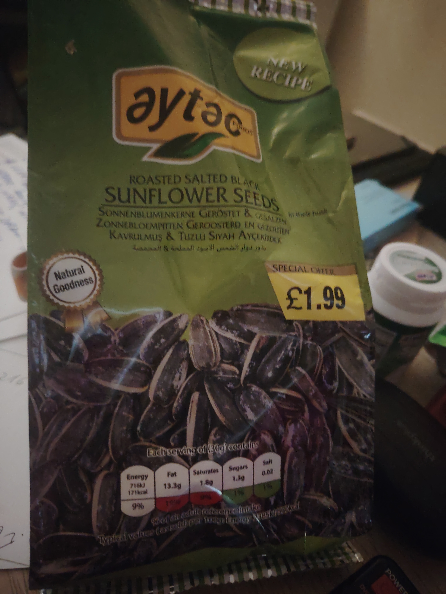 The image shows a close-up of a bag of Aytaç Foods "Roasted Salted Black Sunflower Seeds". The bag is predominantly green with yellow accents. Prominently displayed on the bag is the brand logo "aytaç Foods" and the product name "ROASTED SALTED BLACK SUNFLOWER SEEDS". There is also a "NEW RECIPE" badge and a "SPECIAL OFFER" sticker with a price of "£1.99". Below the product name, there are descriptions in multiple languages including German and Turkish, and possibly Arabic. The bottom half of the bag features a large photograph of sunflower seeds. A nutritional information chart is visible on the lower right side of the bag, indicating values for Energy, Fat, Saturates, Sugars, and Salt per serving. A circular "Natural Goodness" badge is on the left side of the bag. The bag is placed on a surface, likely a table, and the background is blurred but suggests an indoor environment. The overall lighting is artificial, possibly from a lamp or overhead lights. There is no indication of the specific location within Huntingdonshire, United Kingdom.