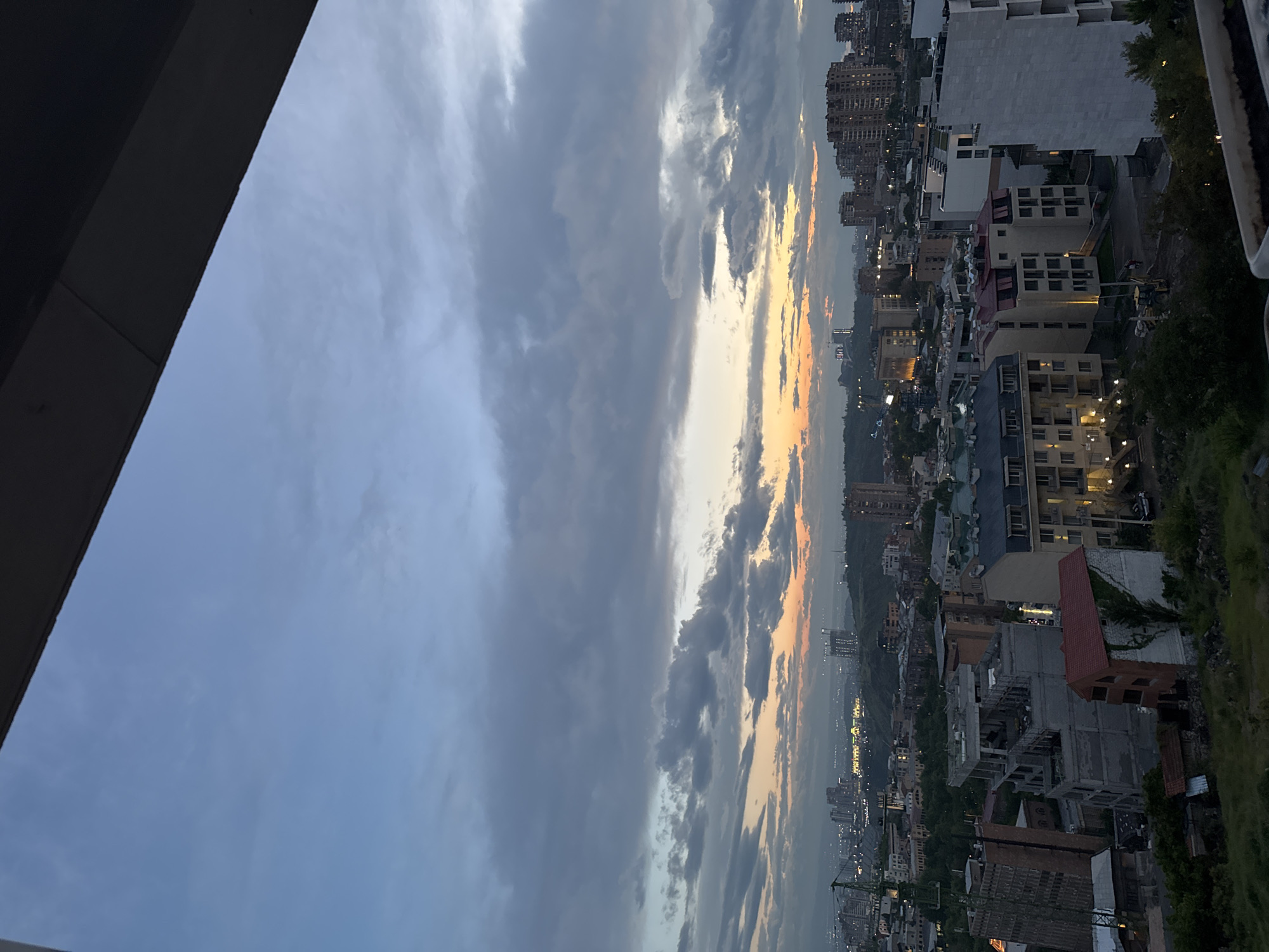 The image depicts a cityscape under a cloudy sky with a hint of sunset. The scene is likely in Yerevan, Armenia, based on the architecture and urban layout. The image is taken from a high vantage point, possibly a balcony or rooftop, overlooking a dense urban area. The architecture is varied, with buildings ranging from modern high-rises to older, low-rise structures. Construction sites are visible amidst the built environment. The sky is predominantly covered with clouds. A strip of orange and yellow light suggests the presence of a sunset or the afterglow of the sun below the horizon. No people are visible in the image.