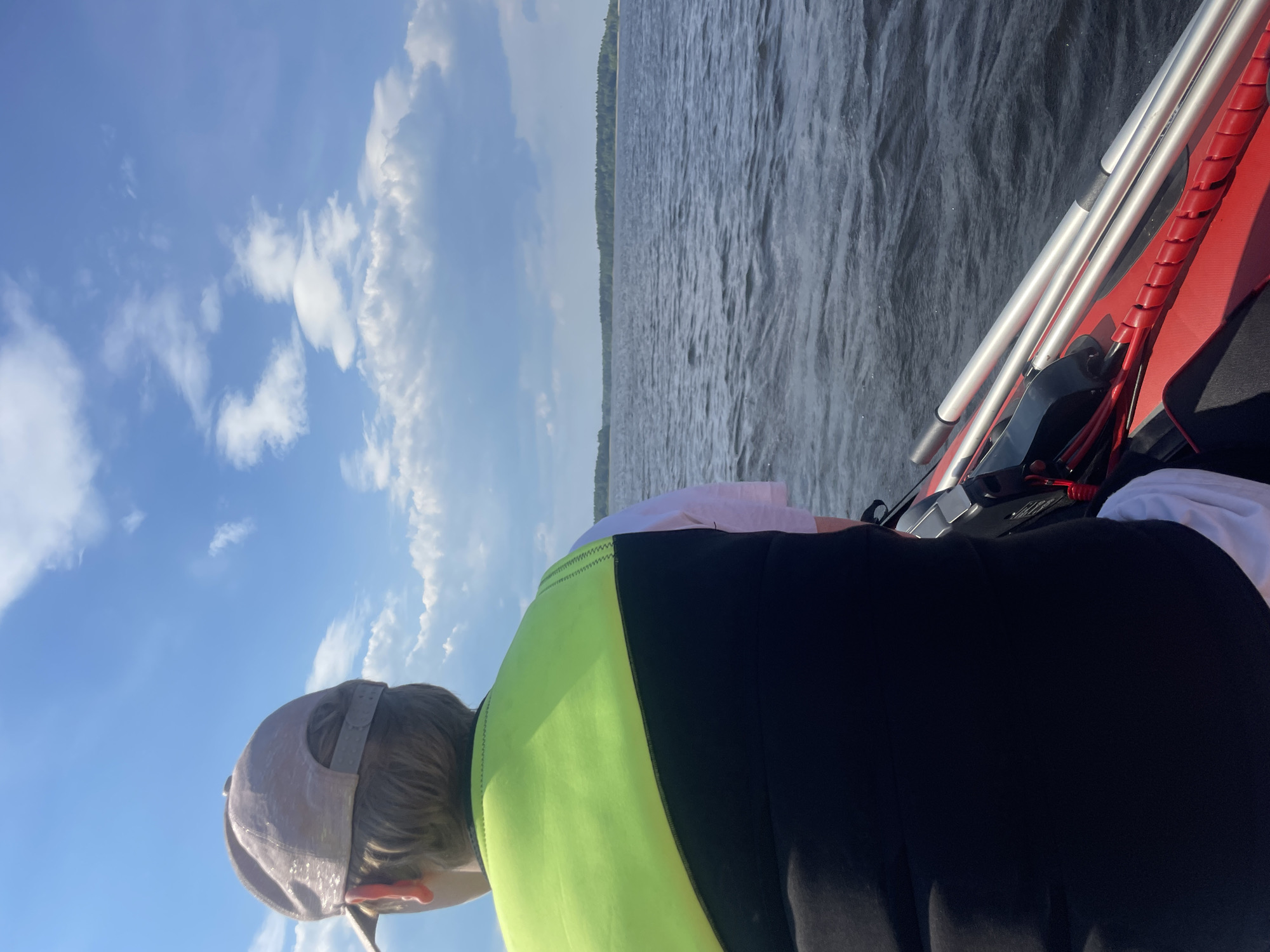 A rear-view, high-angle photograph captures an individual seated in a red inflatable boat on a body of water near Priozersk, Russia. The person wears a light-colored, textured baseball cap over light brown hair and a two-tone life vest, featuring a neon yellow-green upper section and a black lower section, over a white short-sleeved shirt. The individual faces away from the camera, looking towards a distant, flat forested shoreline across the horizon. The water surface is dark blue-grey with visible ripples. Two silver-colored, telescopic poles or oars are laid on the boat's right side, adjacent to a red coiled protective wrap. The upper portion of the scene displays a bright blue sky with scattered white cumulus clouds.