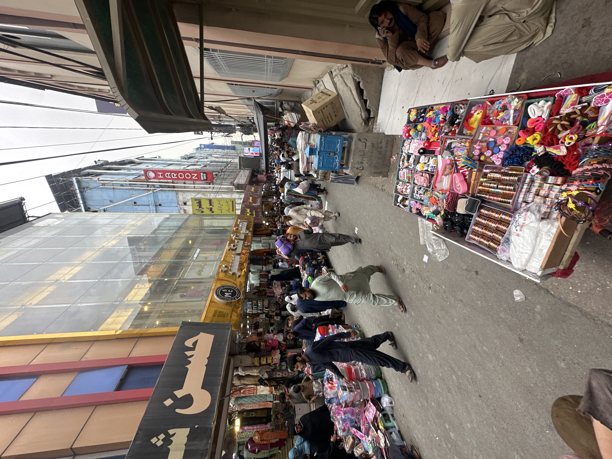 The image shows a crowded street in Quetta City Tehsil, Pakistan. People are walking along a narrow street lined with shops. The street is filled with pedestrians, creating a bustling atmosphere. Shops display various goods, including clothing, accessories, and general merchandise. Signs in Urdu and English adorn the storefronts. Street vendors display their wares along the pavement. The buildings on either side of the street are a mix of old and new construction. Utility poles and overhead wires are visible above the street. A person is sitting on the pavement, talking on a mobile phone.