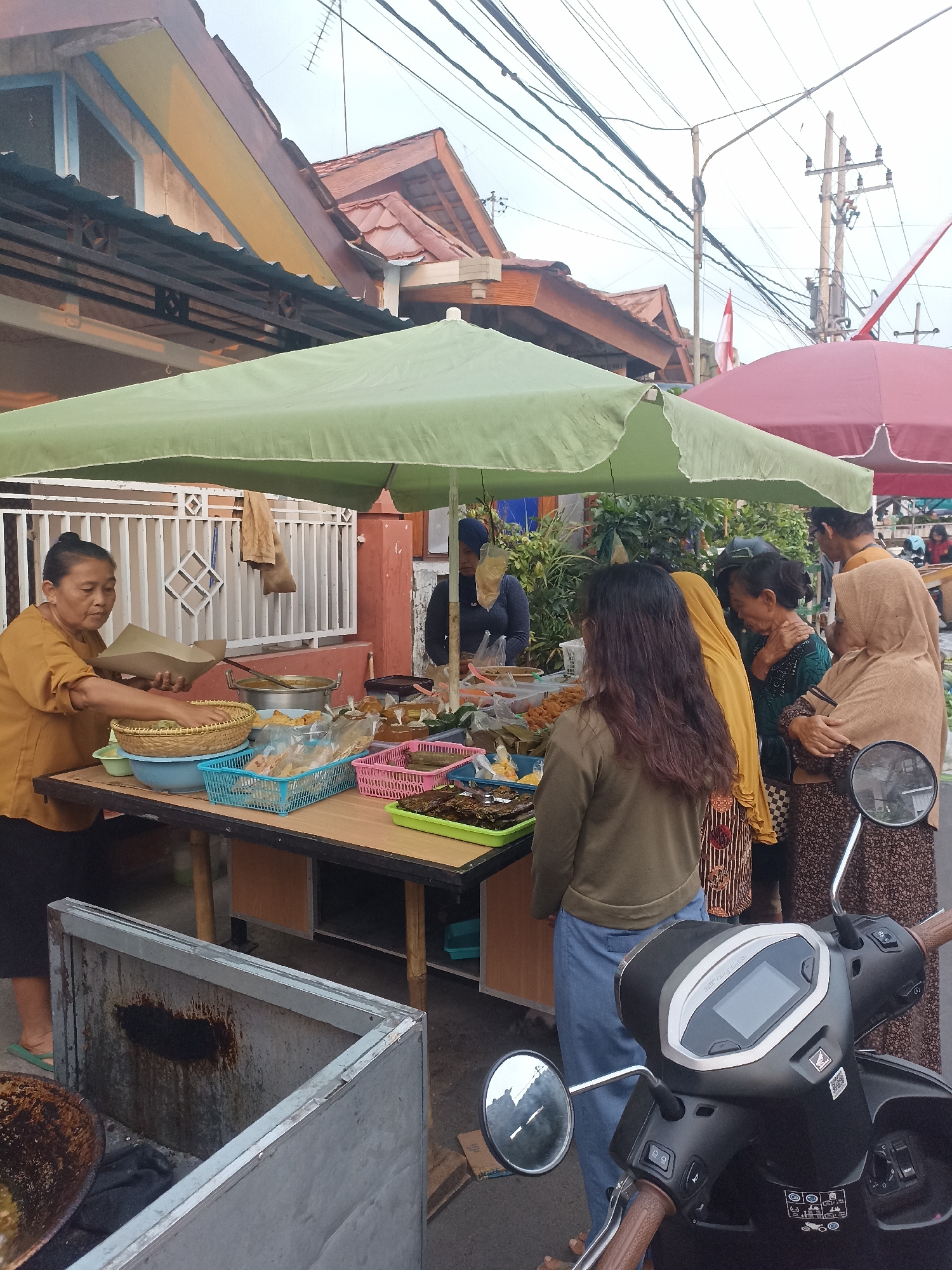 In Mojopanggung, Indonesia, a street vendor is preparing food for customers at a market stall. A woman in a mustard-colored shirt is serving food in paper wrappers to a customer. The stall is covered by a green umbrella and displays various food items in baskets and plastic containers. Several customers are gathered around the stall, some looking at the food and others interacting with the vendor. A motorcycle is parked in the foreground, and buildings with tiled roofs line the street in the background. The scene depicts a bustling market with a focus on food sales.