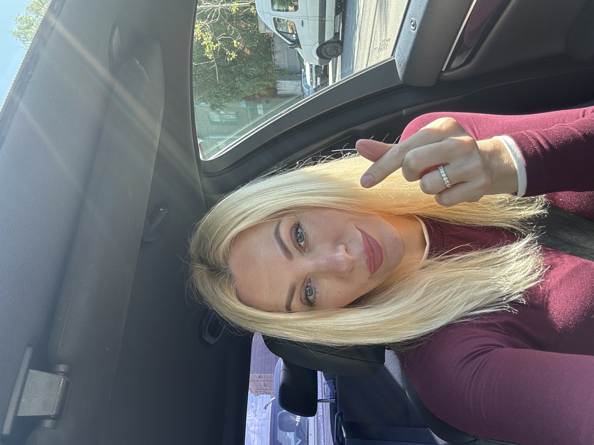 A woman with blonde hair and blue eyes is taking a selfie inside a car. She is wearing a maroon long-sleeved shirt and a ring on her left hand. She is making a heart shape with her fingers. The car's interior is visible, including the ceiling, dashboard, and passenger seat. Through the car window, a street scene in Saint Petersburg, Russia is visible, with buildings, trees, and a white van. The sun is shining, casting a warm glow on the scene.