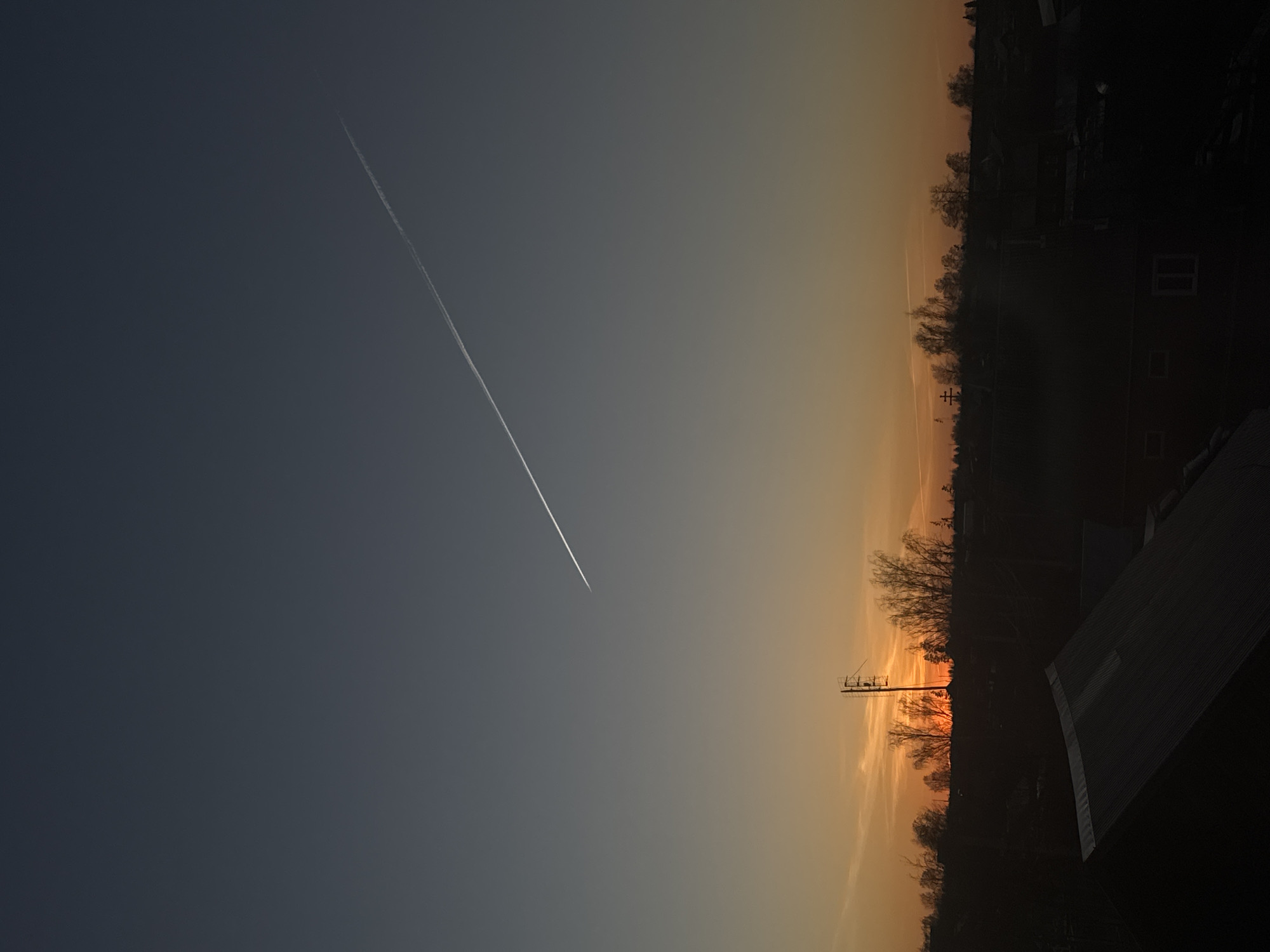 The image depicts a sunset view in Фоки, Russia. The sky transitions from a dark blue-gray on the left to an orange glow on the right, indicating the direction of the sunset. A contrail is visible in the upper left portion of the sky. The horizon is lined with dark silhouettes of trees and buildings, with the suggestion of residential structures. A utility pole stands out against the sunset. A section of a roof is visible in the lower right foreground.