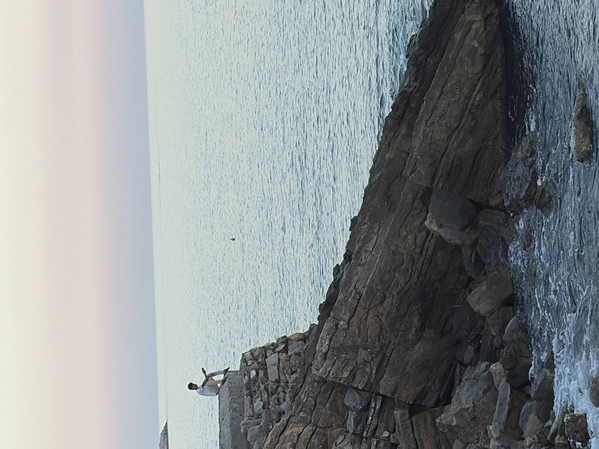 In a dramatic coastal scene, a lone person is perched precariously on a rocky outcrop overlooking the sea. The individual, wearing a light-colored shirt and darker trousers, is seated with their legs drawn up, facing away from the viewer and towards the vast expanse of the water. The rough, textured surface of the dark grey rocks contrasts sharply with the smooth, undulating appearance of the sea, which shimmers with a pale blue hue, suggesting calm waters.
The lighting in the image indicates it is likely either early morning or late afternoon, with a soft, diffused light bathing the scene. The sky transitions from a pale, almost white tone on the left to a more vibrant blue further out. There are no clear indications of specific weather conditions, but the sea appears placid. The location is identified as Imperia, Italy, suggesting a Mediterranean coastline with its characteristic rugged cliffs and clear waters. A small, reddish object is visible in the middle distance over the water, though its nature is unclear. The overall mood is one of solitude and contemplation, with the person seemingly absorbed in the view. No text is visible in the image.