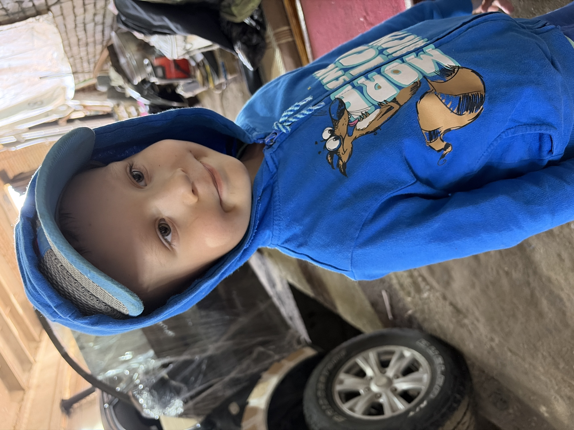 In Chaikovsky, Russia, a young boy wearing a blue hooded jacket and a baseball cap poses for a photo. He stands in what appears to be a garage or workshop, with a black vehicle partially visible behind him. The environment is cluttered with various objects, including what looks like a tire and other unidentifiable items. The boy's expression is slightly amused, with a hint of a smile.