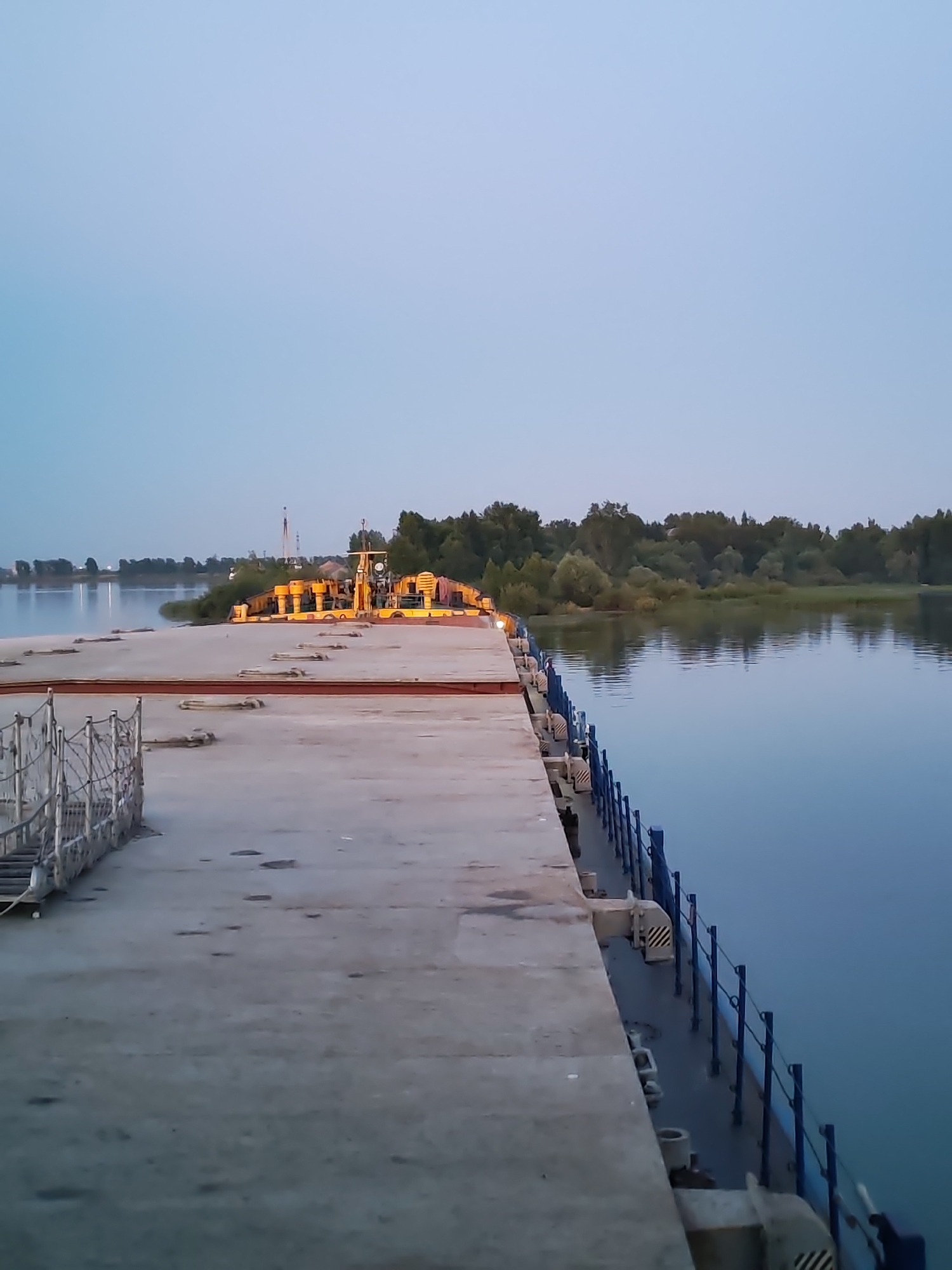 The image depicts a concrete dock extending into a calm body of water. On the dock, a yellow barge with industrial equipment is visible. The barge is illuminated by several lights, suggesting it might be operational or undergoing work. The water reflects the twilight sky, which is a pale blue. A line of trees and foliage borders the opposite bank of the water, with some structures and distant lights indicating a settlement. The scene is set in the city of Bol'shie Mkhi, Russia.