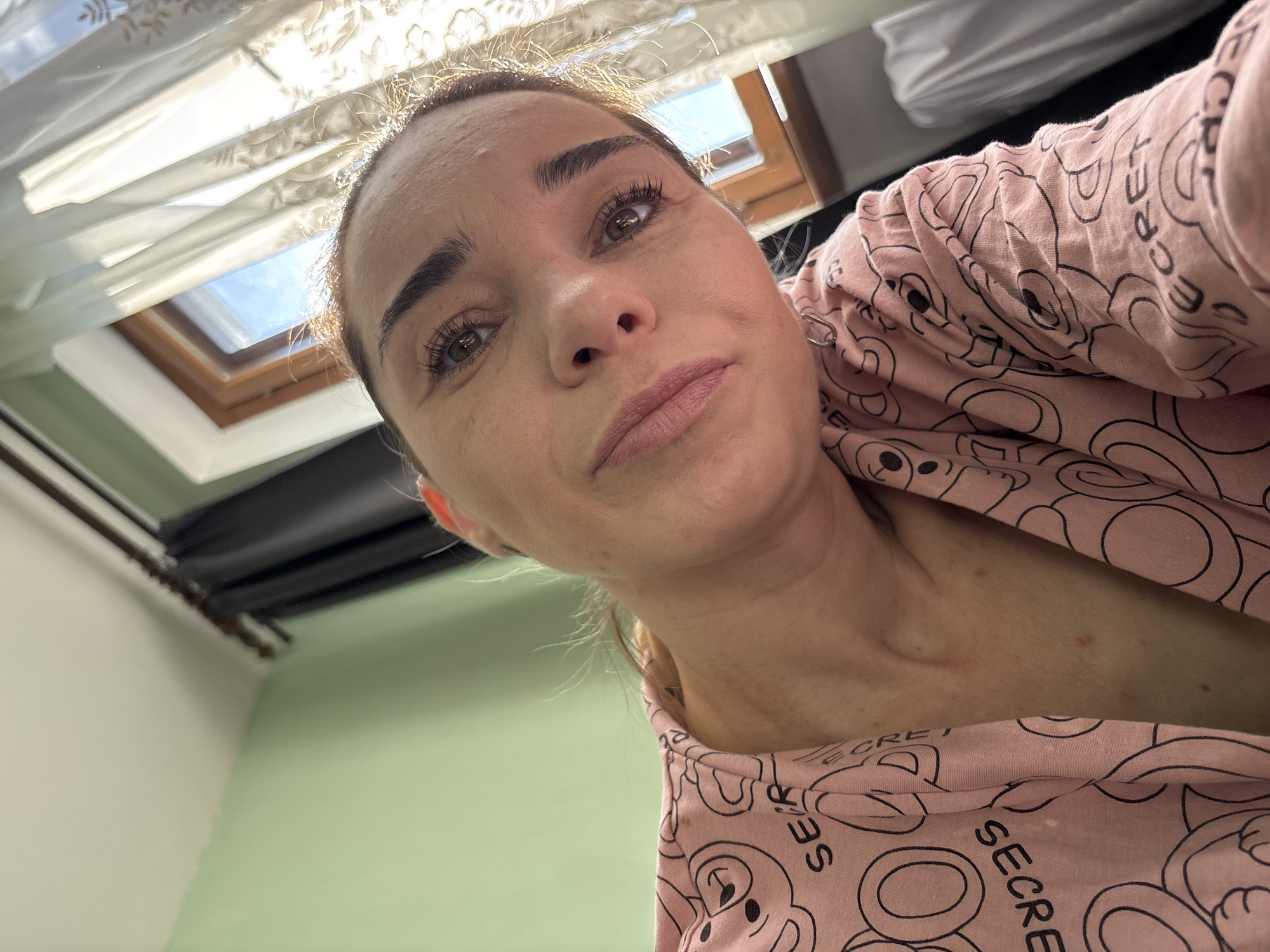 A woman is captured in a close-up, low-angle selfie. She is wearing a pink garment with a repeating pattern of teddy bears and the word "SECRET." The background reveals a room with a pale green wall and a window. Sunlight streams through sheer, embroidered curtains, illuminating the scene. A dark curtain is visible on the left side of the window. The woman's face is the primary focus, with her gaze directed slightly away from the camera. Her hair is pulled back, and she has makeup on, including mascara and lipstick. The overall impression is of an indoor setting, possibly a bedroom, in what could be a residential area. The provided information does not specify the city or country.