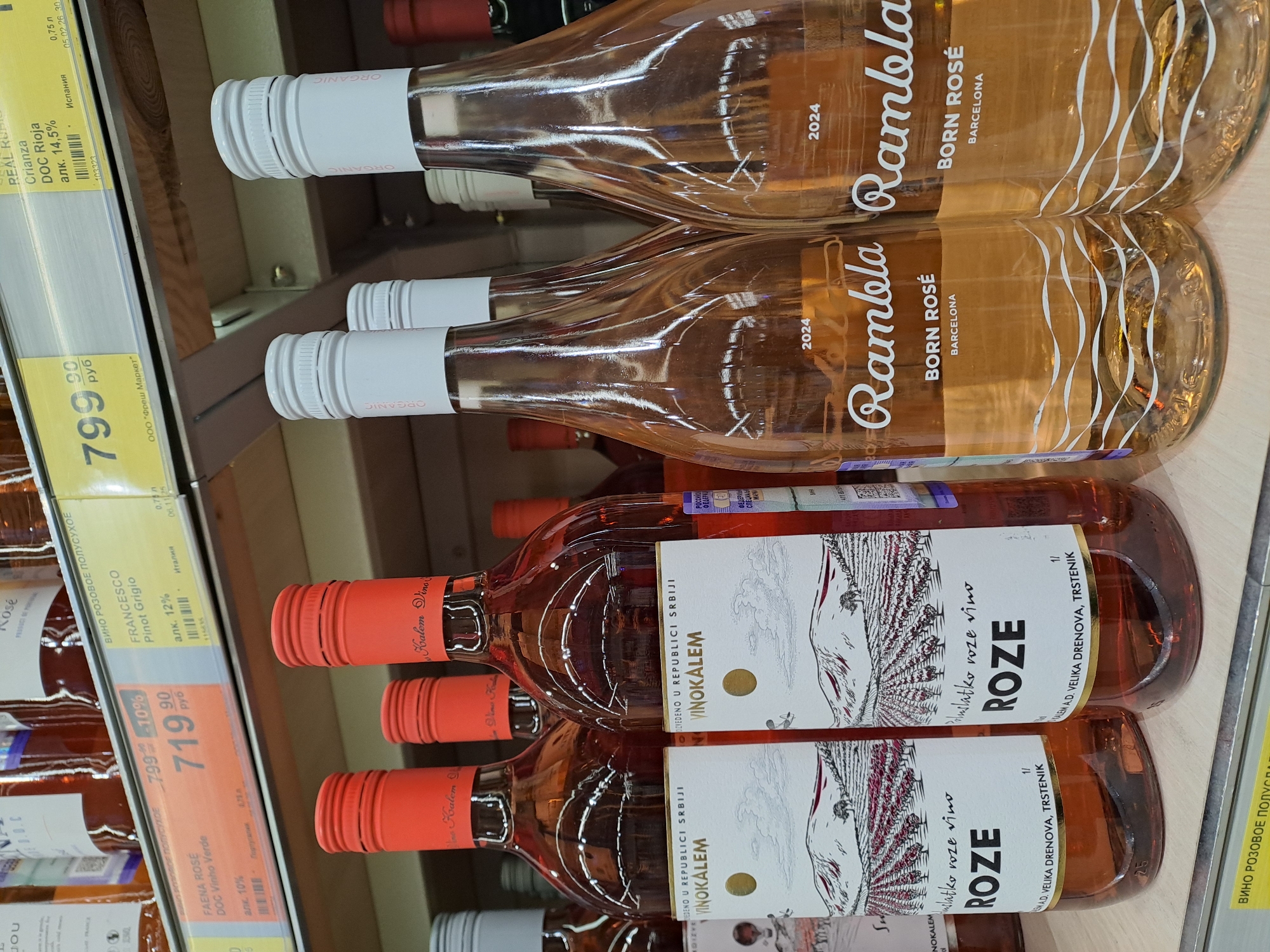 This image shows a shelf in a store displaying several bottles of rosé wine. The top two bottles are "Rambla Born Rosé Barcelona 2024". Below are bottles of "Vinokalem ROZE", which are from Serbia. Price tags are also visible, one listing "FRANCESCO Pinot Grigio" at 719.90 rubles and another listing "REAL Rioja" at 799.90 rubles. The store appears to be in Elektrostal, Russia due to the price tags and Cyrillic script on the labels.