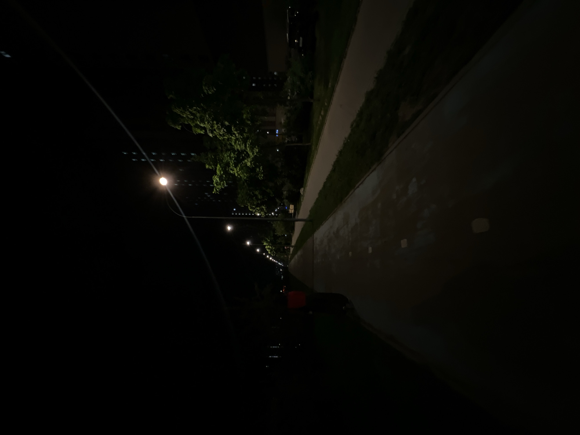 The image depicts a night scene in Moscow, Russia. A row of streetlights illuminates a pathway lined with trees and buildings. The pathway appears to be adjacent to a wall or barrier. The area is mostly dark, with the streetlights providing the primary source of illumination. There is a red object in the middle distance.