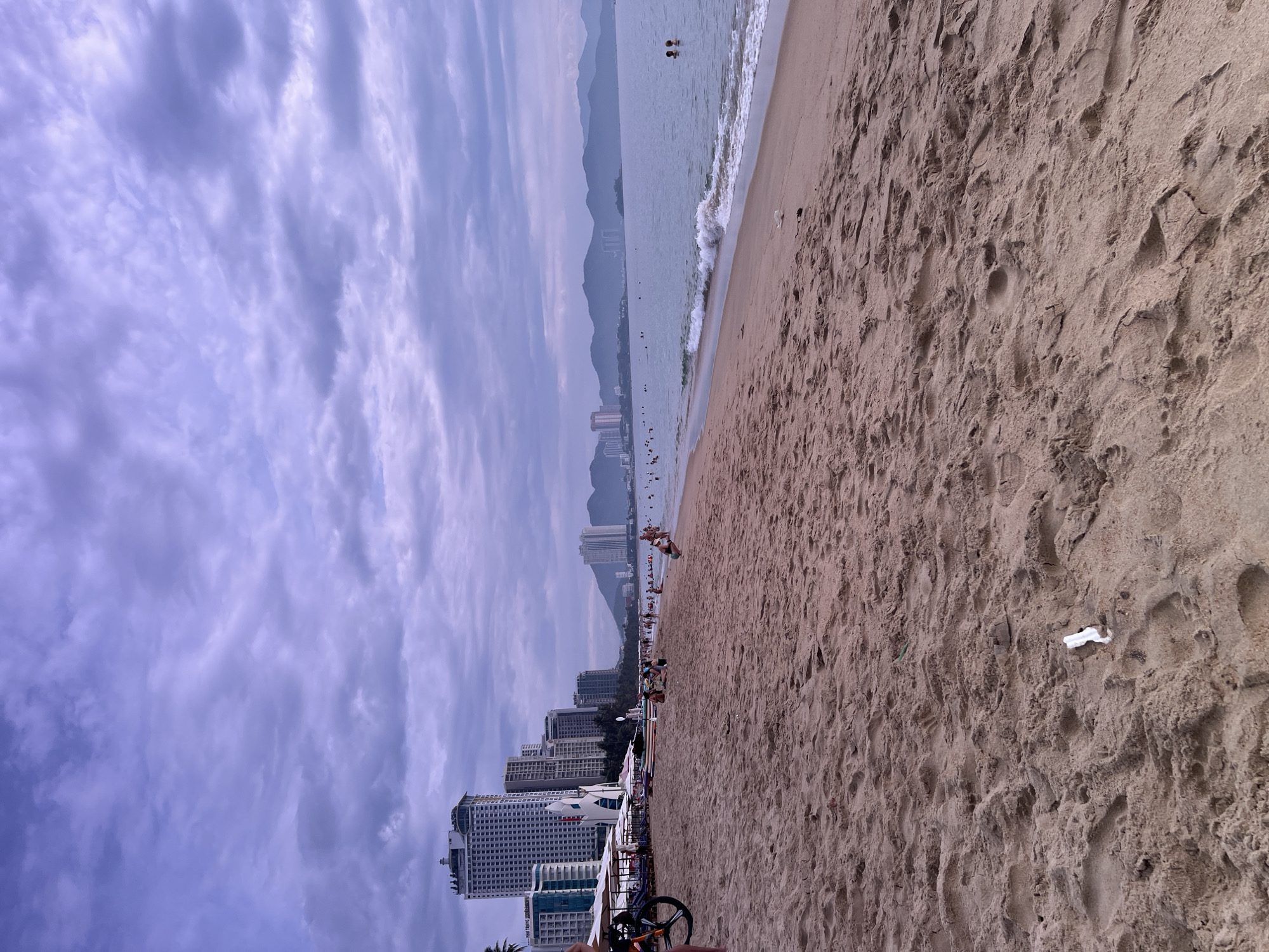 This image depicts a wide, sandy beach with the ocean on the right and a cityscape in the background. The sky is overcast with a heavy cloud cover. In the foreground, the sand is textured with footprints and some debris, including a crumpled white object.
Along the shoreline, there are people scattered across the beach, some swimming in the shallow water, while others are walking or sitting. Beach umbrellas and lounge chairs are visible, suggesting a popular tourist destination.
Dominating the background on the left side of the image is a cluster of tall, modern buildings, including a prominent white high-rise with a distinctive curved facade and other multi-story hotels and apartments. Further in the distance, more buildings line the coast, and a mountain range forms the horizon.
The overall scene is characteristic of a busy urban beach in Nha Trang, Vietnam, likely taken during the day, possibly in the late afternoon given the lighting conditions and the muted colors of the sky.