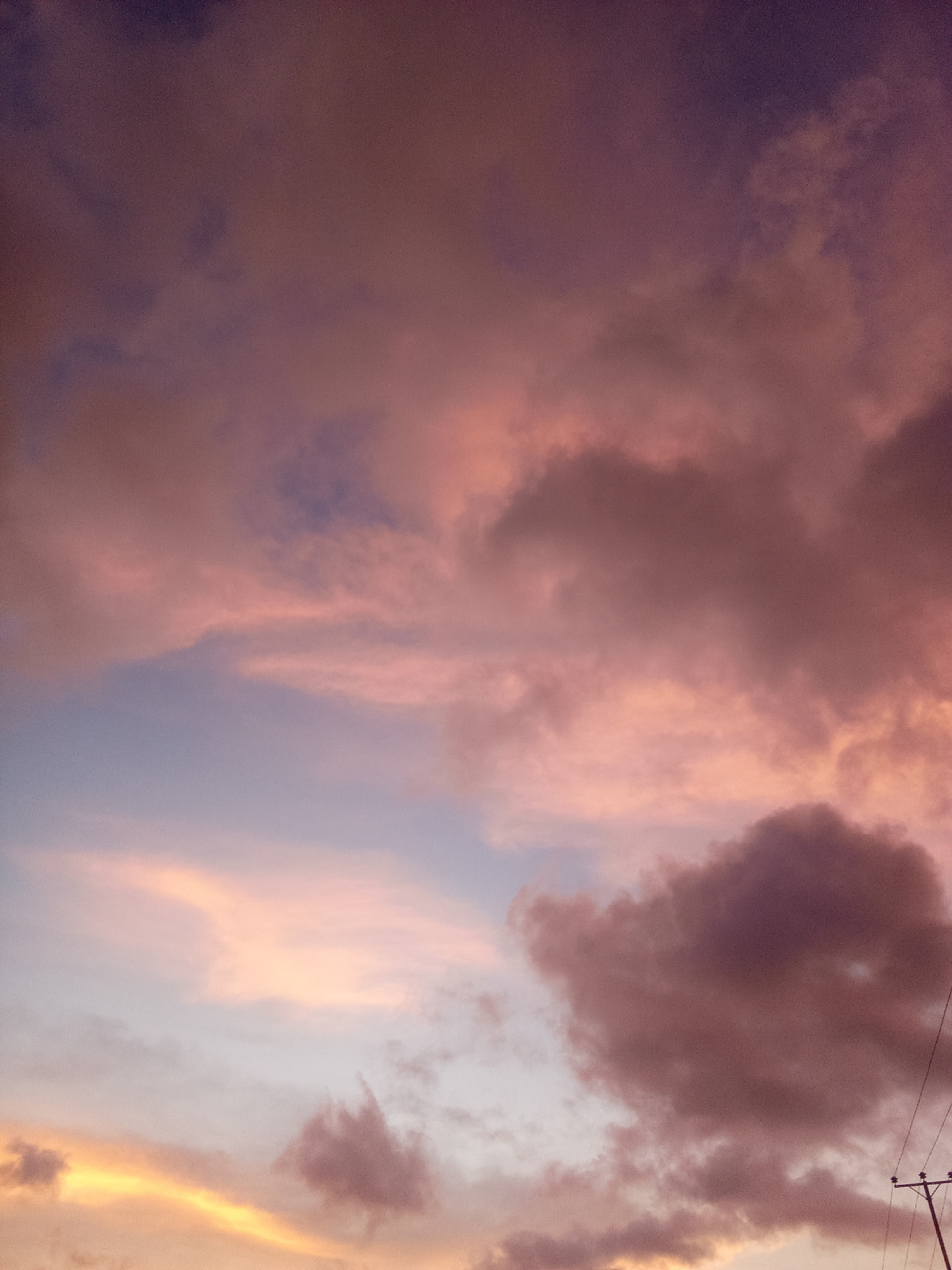 The image shows a dusky, colorful sky. The scene is dominated by a blend of pink, orange, purple, and blue hues, suggesting a sunrise or sunset. Clouds are visible throughout the frame, with varying densities and shades of pink and purple. In the lower right corner, there's a glimpse of a utility pole, hinting that this may be an urban or suburban environment in Juan Griego, Venezuela. The focus is primarily on the sky, with no visible people or specific actions taking place.