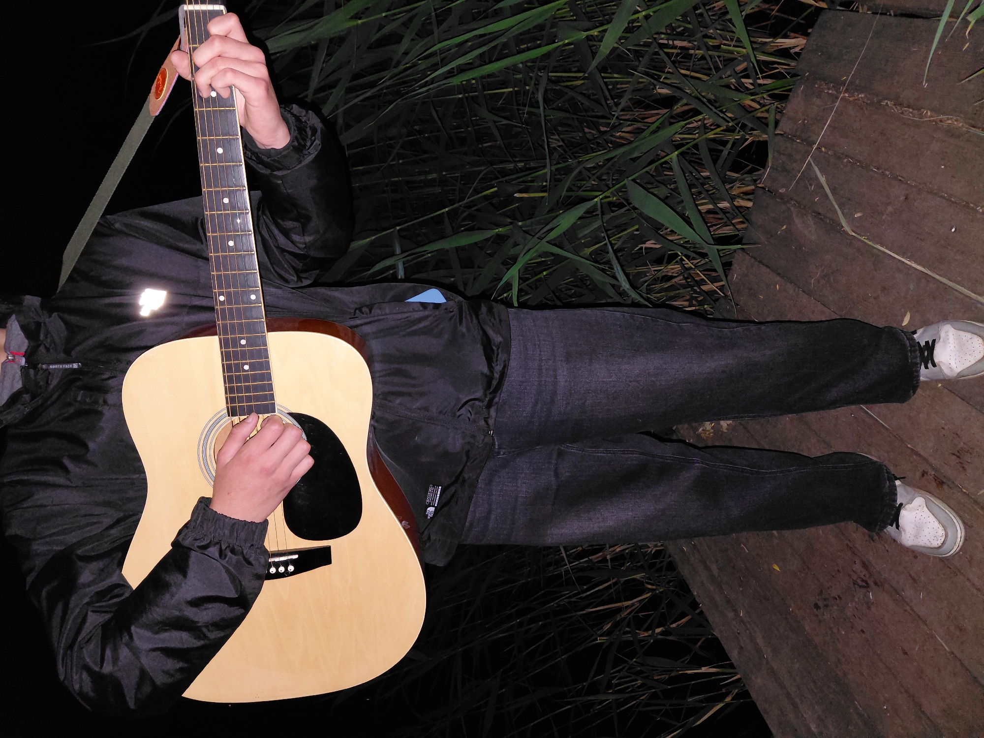 A person is depicted playing an acoustic guitar at night in Ustyvytsia, Ukraine. The image, rotated clockwise, shows the individual from the torso down, standing on a wooden boardwalk or pier. They are wearing a black jacket with a visible "NORTH FACE" tag, dark jeans, and white sneakers. Tall, dark reeds or grass are abundant to the left and behind the person, indicating an outdoor, natural setting, possibly near water.
The subject is actively playing an acoustic guitar with a light wood finish and a black pickguard. Their left hand is positioned on the fretboard, holding down strings, while their right hand is near the soundhole, ready to strum or pluck. A light-colored guitar strap is slung over their left shoulder. The scene is dimly lit, characteristic of nighttime, with some artificial light creating reflections, notably a bright spot on the front of the jacket. The overall impression is one of a musician enjoying their instrument in a tranquil, dark environment.