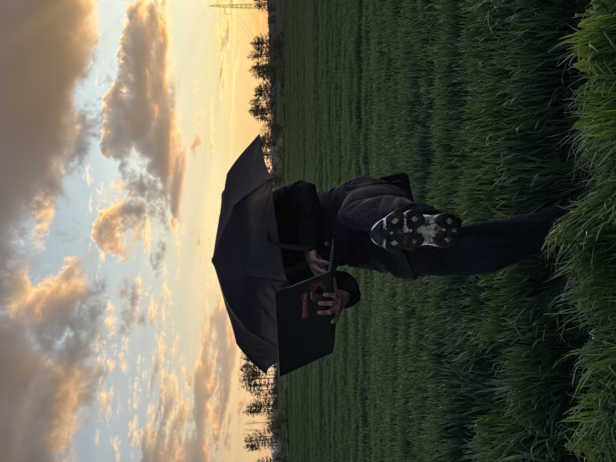 A person lies on their left side in a grassy field under a cloudy sky with a power line in the distance. The person wears dark clothing and sneakers, holding a black laptop computer and a black umbrella. The laptop's logo, "MOBILE FOX" is visible. The field is green with taller grass on the right side of the frame. The location is likely in Shumen, Bulgaria, due to the prompt's request.