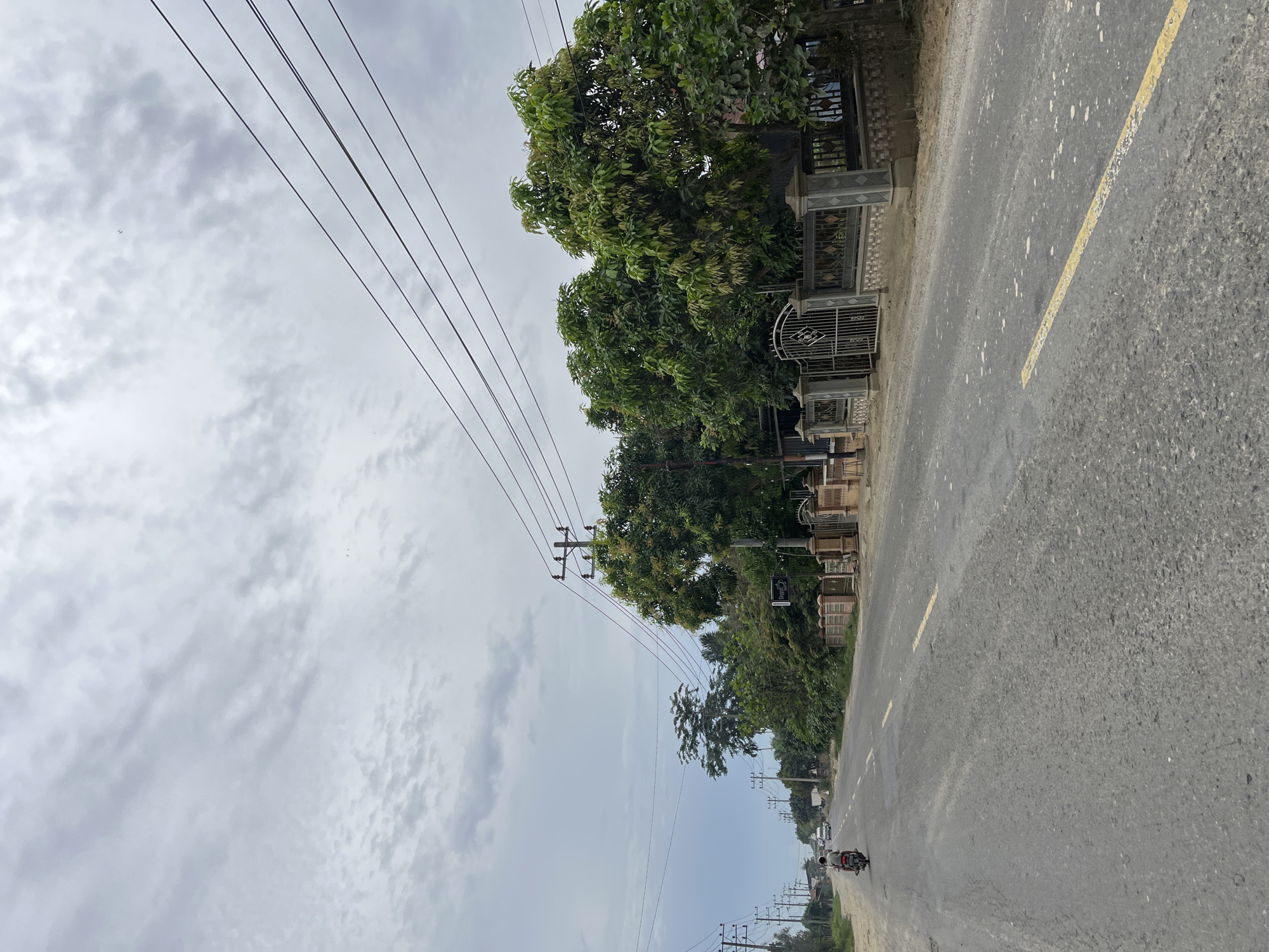 The image depicts a straight, asphalt road with a yellow dividing line, characteristic of roads in Simpangempat, Indonesia. The road is flanked by a dusty shoulder on the left and a low, ornate fence with stone pillars on the right, separating the road from a residential area or property. Lush green trees, some with visible foliage and fruit, line the right side of the road. Overhead, a network of power lines stretches across the sky, supported by utility poles. The sky is overcast with soft, grey clouds.
In the foreground, a motorcycle carrying a single rider is traveling away from the viewer. Further down the road, several cars are visible in the distance, suggesting moderate traffic. The overall scene conveys a sense of a rural or semi-urban environment in Indonesia, with the road serving as a primary transportation artery. The presence of a residential fence and mature trees suggests a developed area within or adjacent to the town.