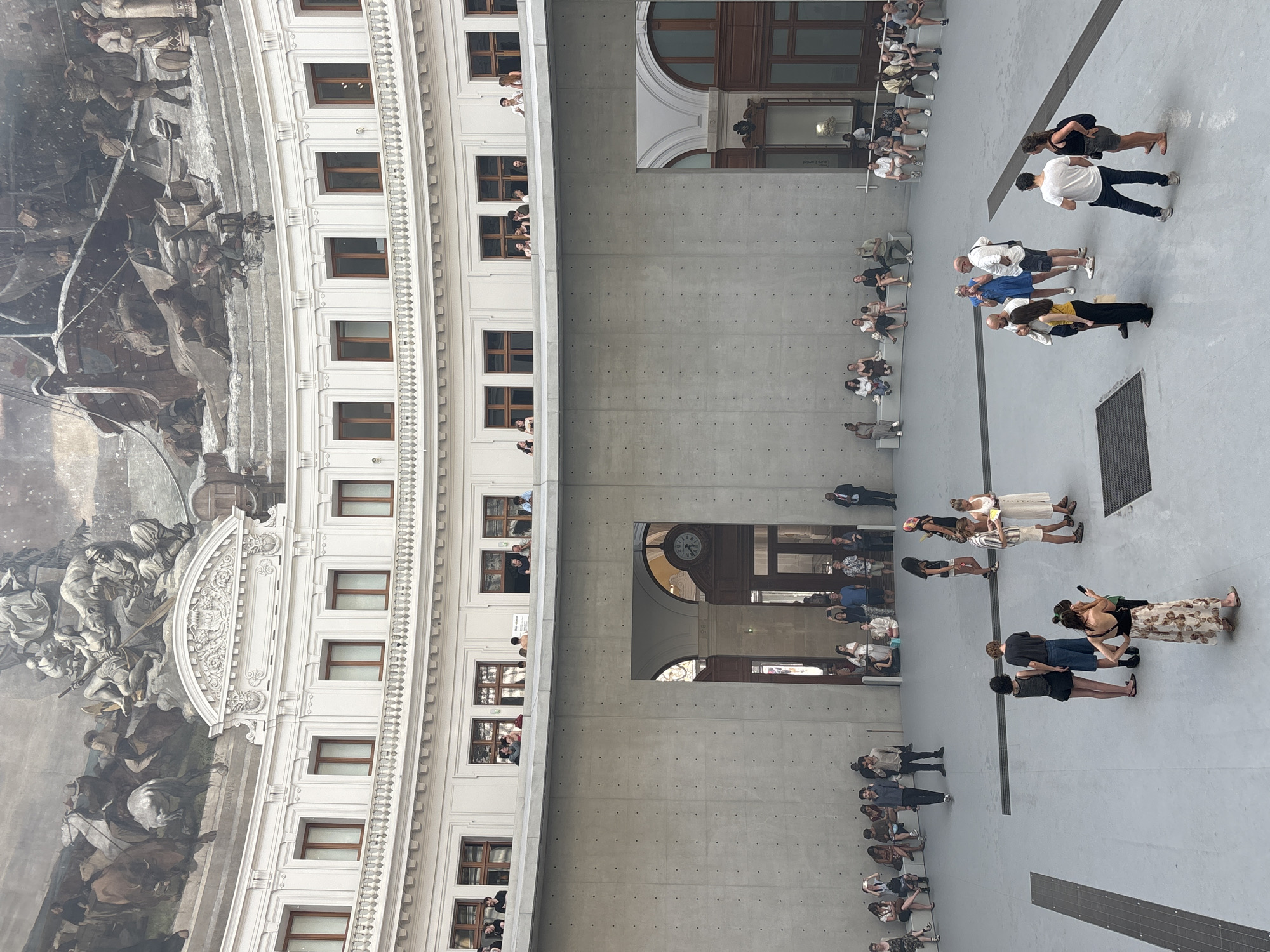The image depicts the interior of the Bourse de Commerce in Paris, France, a circular building featuring a large open central space surrounded by multiple levels. The upper level features ornate white architecture with arched windows and a large fresco ceiling depicting maritime scenes. People are visible on this upper level, looking out over the central area.
The central space has a high, unfinished concrete wall with a large arched opening leading to an interior with a clock and more classical architecture. In the center of this space, a group of people are gathered. Some are standing and conversing, while others are seated on benches along the concrete wall, observing the scene or interacting with each other. The floor is a light grey concrete. The overall atmosphere suggests a public space, possibly a museum or exhibition hall, with visitors present.