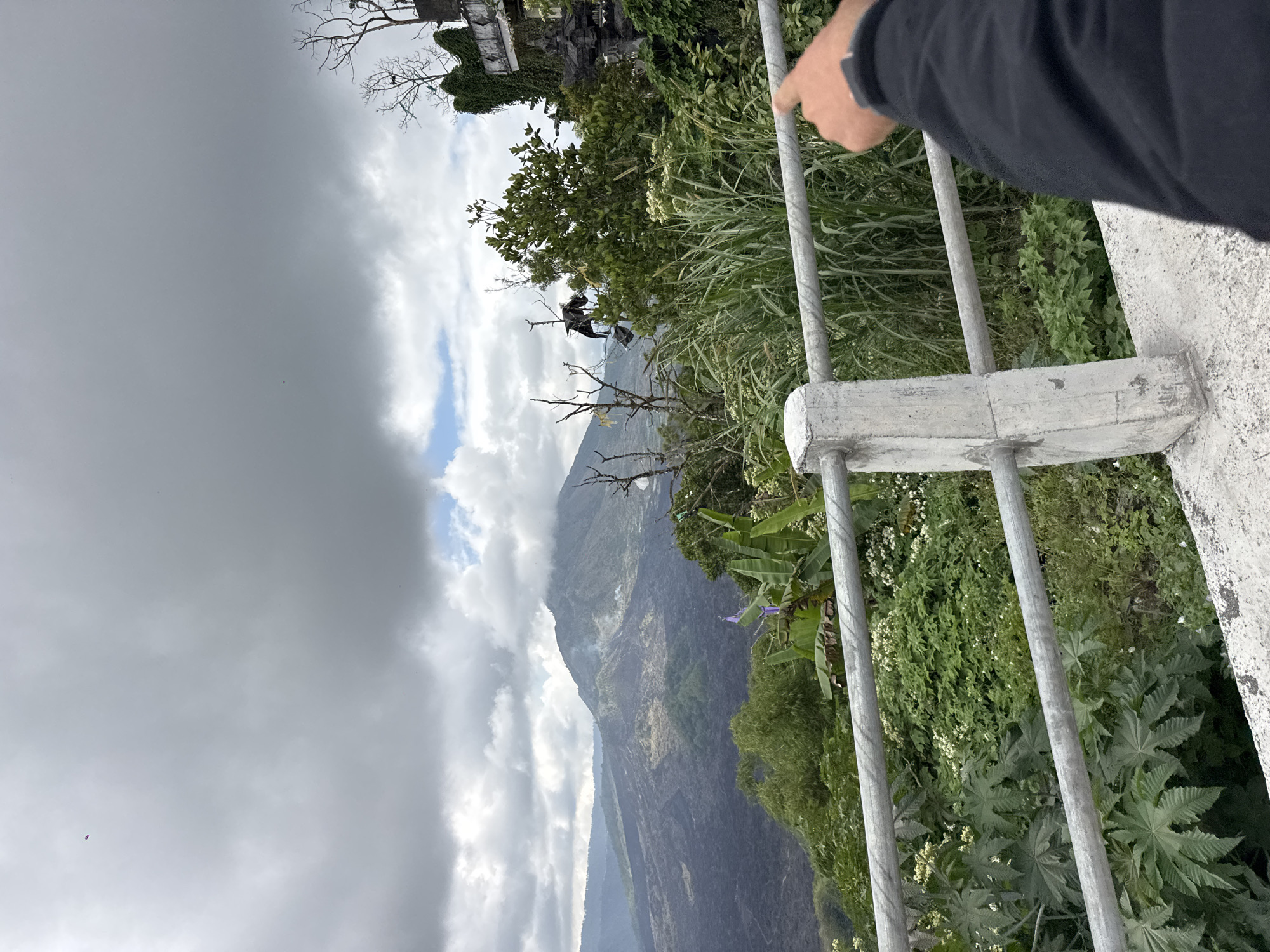 The image depicts a view of Mount Batur in Kintamani, Bali, Indonesia. In the foreground, a hand wearing a black jacket points towards the mountainous landscape. A metal railing with concrete posts runs horizontally across the lower portion of the image, separating the viewer from the lush green vegetation and the volcanic terrain beyond. Mount Batur, a stratovolcano, is visible in the background, partially obscured by clouds. The sky is overcast with a mix of gray clouds and hints of blue. The landscape is characterized by the dark, rocky slopes of the volcano, which appear to be solidified lava flows, contrasting with the vibrant green foliage. A small, distant object, possibly a paraglider or a bird, is visible in the sky in the upper left quadrant of the image.
