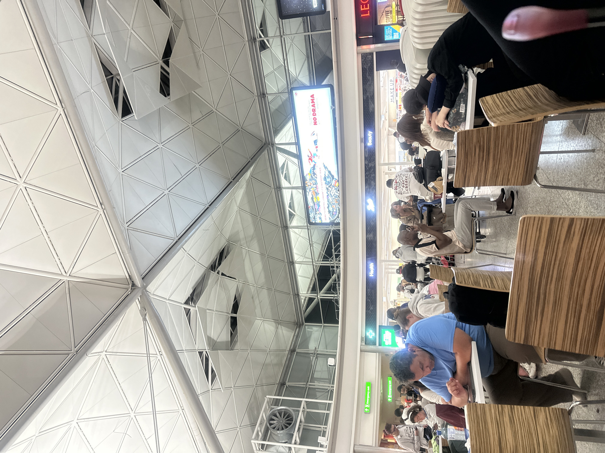 The image depicts an interior scene at Stansted Airport in Uttlesford, United Kingdom. The architecture features a distinctive geometric ceiling with white panels and metal supports. In the foreground, several individuals are seated at tables, some engaged with electronic devices, others conversing, and one person is asleep. Various individuals are visible in the background, indicating a busy public space. Signage for "Health," "Beauty," and a "Coffee" establishment are discernible. A large digital billboard displays an advertisement with the text "NO DRAMA" and "GOING TO CENTRAL LONDON IS FASTER BY TRAIN." A large industrial fan is mounted on a metal structure to the left. The overall environment appears to be a food court or seating area within an airport terminal.
