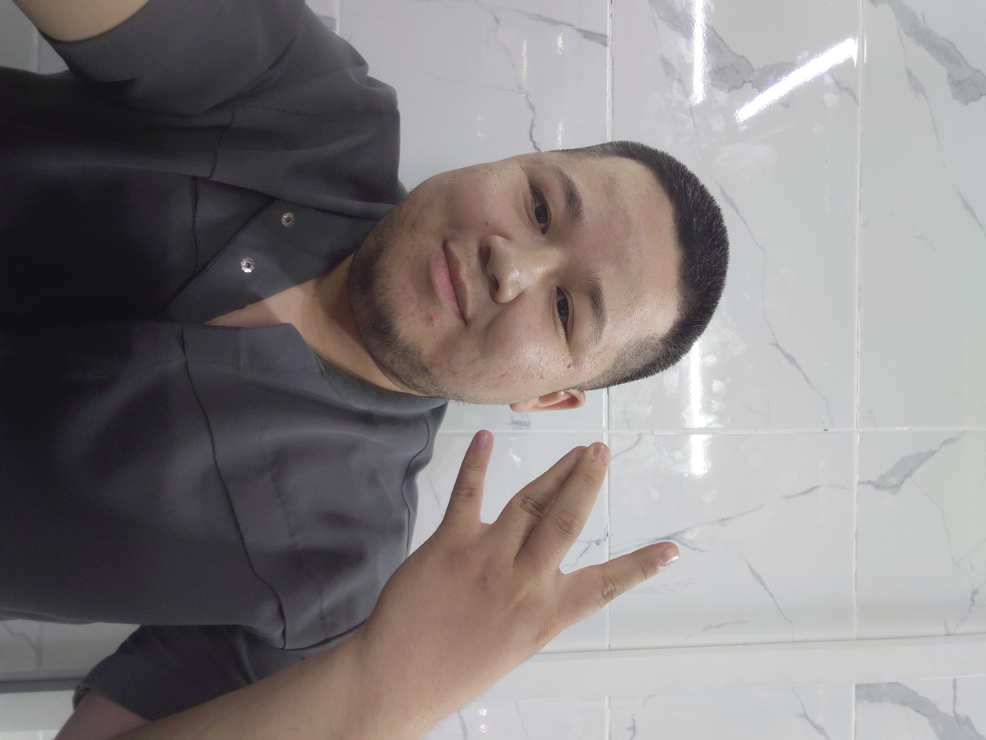 In a selfie taken indoors, a man with a short haircut and stubble is centered in the frame. He is wearing a dark gray collared shirt with visible snaps. The background consists of white tiled walls with marble-like veining. The man is making a hand gesture with his right hand, with his index and middle fingers extended and his ring and pinky fingers curled. The lighting in the scene is bright, creating reflections on the glossy tiles. The image does not contain information that allows for identifying a specific location within Aktau, Kazakhstan.