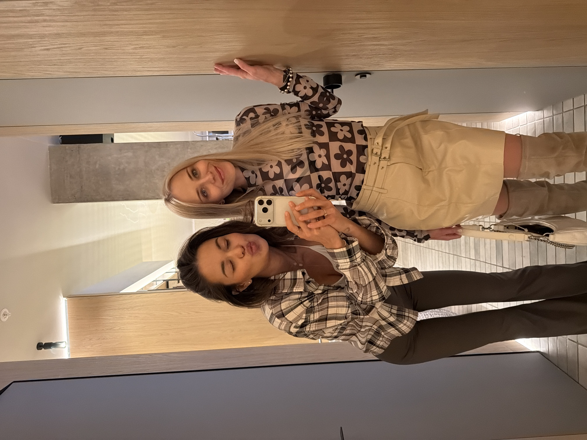 Two women are taking a selfie in a dressing room. The woman on the left is wearing a plaid shirt and dark pants, and she is making a kissing face. The woman on the right is wearing a brown and black floral top, a tan skirt with belts, and tan boots. She is holding the phone. The dressing room has wooden walls and a gray floor. It is unknown whether this image was taken in Krasnoyarsk, Russia.