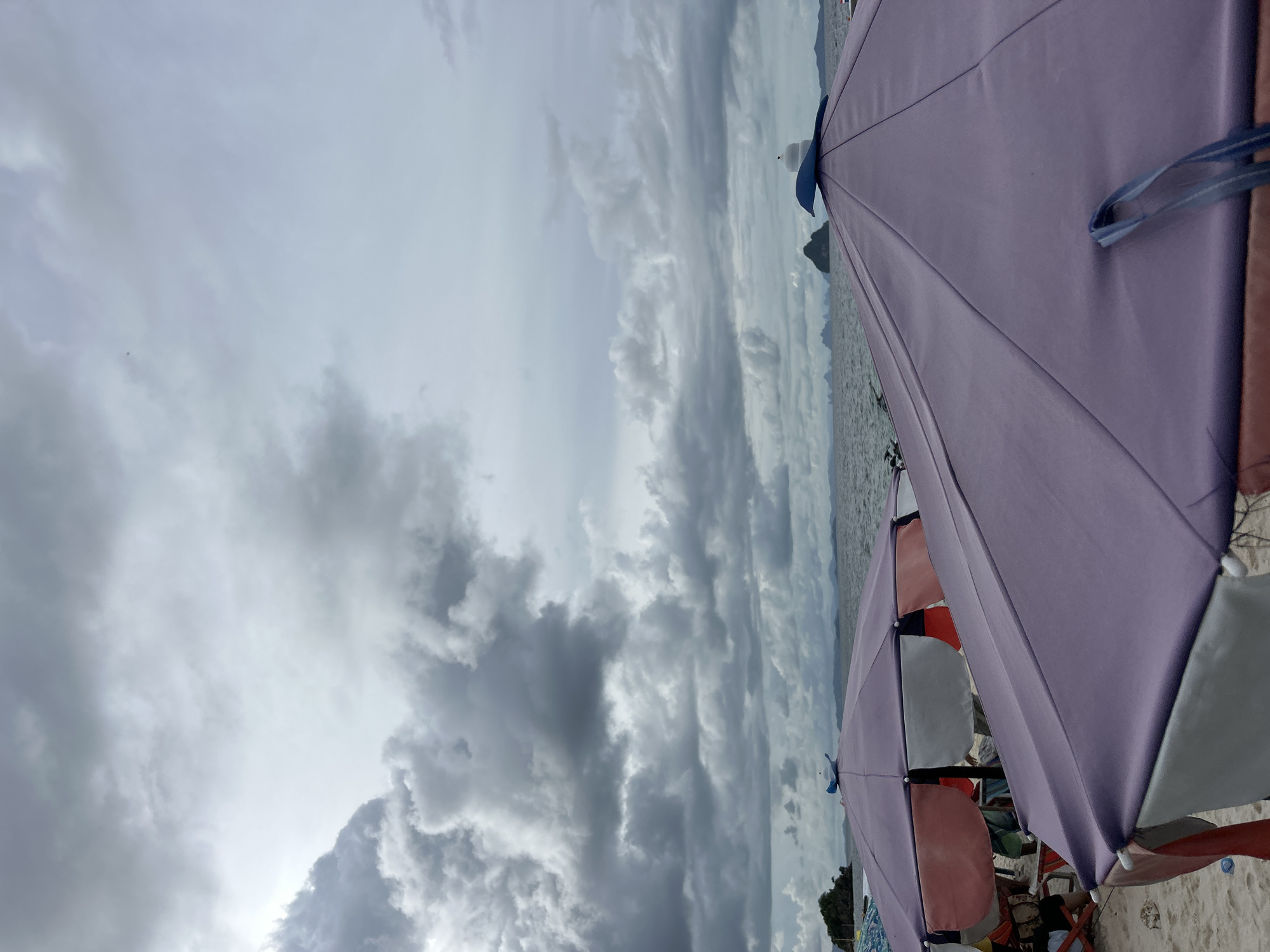 The image depicts a beach scene with several beach umbrellas open under a cloudy sky. The umbrellas are in various colors, including purple, pink, and white. Some people are visible beneath the umbrellas, suggesting they are enjoying a day at the beach. The sea is visible in the background, with a distant island or landmass on the horizon. The sky is filled with dramatic, dark clouds, indicating overcast or possibly stormy weather. The overall impression is that of a beach day interrupted or characterized by inclement weather, yet people are still present and utilizing the beach amenities. The location is identified as Tambon Phru Nai, Thailand, which is a coastal area. No text is visible in the image. The lighting suggests it is daytime, but the heavy cloud cover obscures direct sunlight.