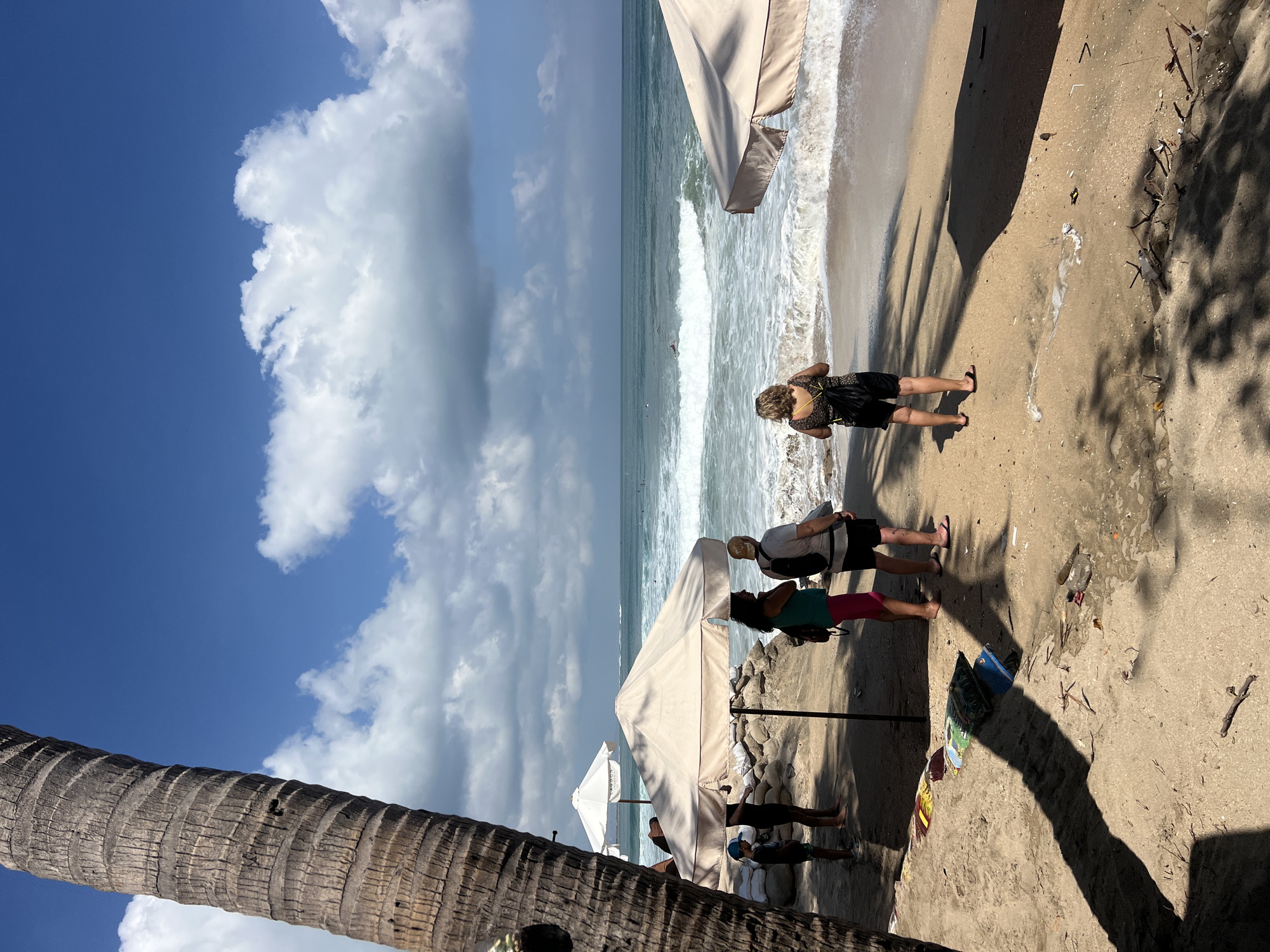 The image shows a beach scene in Kuta, Indonesia. In the foreground, a large palm tree trunk is visible on the left side. Several people are standing on the sandy beach, some looking out at the ocean, while others are positioned under beach umbrellas. One person is wearing a green top and pink pants, another a white shirt and black shorts, and a third a black and yellow patterned top with black shorts. A child in a blue hat is also present. Beach umbrellas, some open and some closed, are scattered across the sand. In the background, the ocean waves are crashing on the shore under a blue sky with scattered clouds. There are also sandbags visible, likely for coastal protection. The overall impression is a relaxed beach day with a few individuals enjoying the coastal environment.