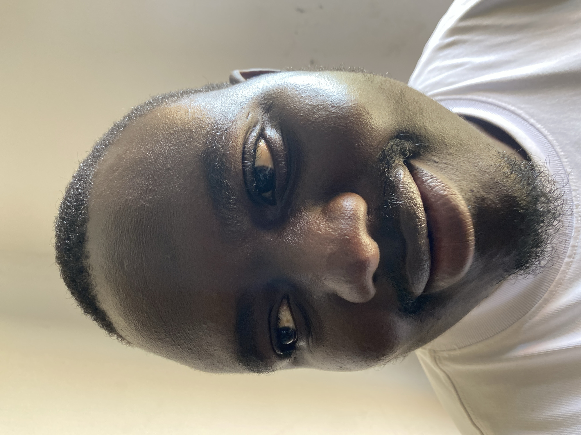 The image is a close-up, low-angle, distorted portrait of a Black man with dark skin, a receding hairline, and short, dark hair. He has a prominent forehead, a wide nose, and a mustache and goatee. His right eye is visible and looking slightly to the left, while his left eye is mostly obscured. He is wearing a white t-shirt with a ribbed neckline. The background is a plain, light-colored wall, possibly illuminated by natural light from the left. The distortion suggests a wide-angle lens was used. The image appears to be a selfie taken indoors. There is no visible indication of the specific location in Jos, Nigeria.