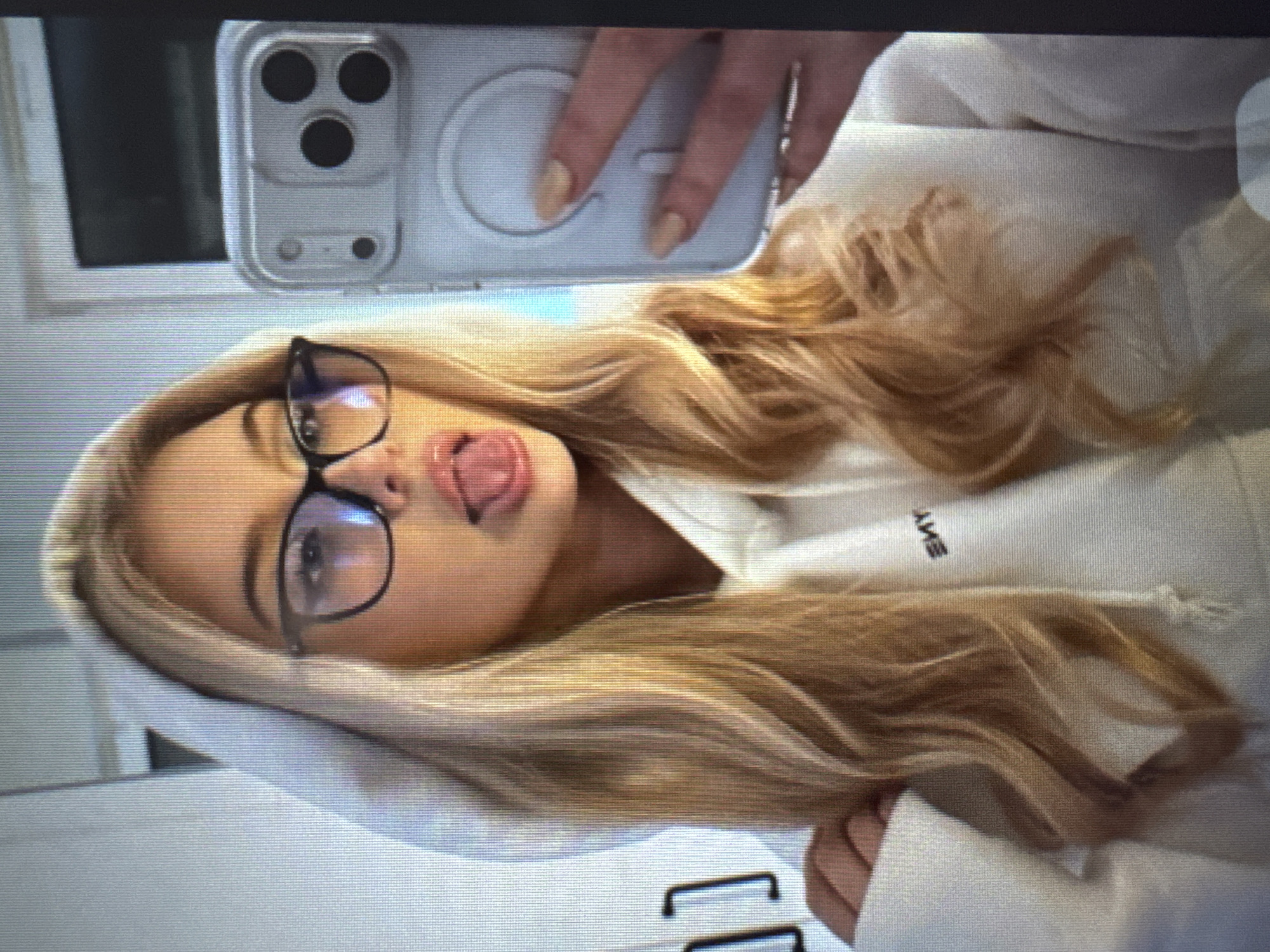 The image displays a headshot and upper torso of a young woman, captured in a selfie. The woman has long blonde hair and wears glasses with dark frames. She is dressed in a white hooded sweatshirt. Her expression is playful, with her tongue extended. She is holding a clear-cased smartphone, which appears to be an iPhone due to the distinct camera module and MagSafe ring on the back. Her fingers, featuring light-colored manicured nails, are visible gripping the device. The background is mostly out of focus, consisting of light tones and what might be a window or light-colored wall in the upper left. Visible on her sweatshirt are the mirrored letters "SMY", suggesting a word like "YMS" or part of a brand name. The overall scene conveys a casual, lighthearted moment.
