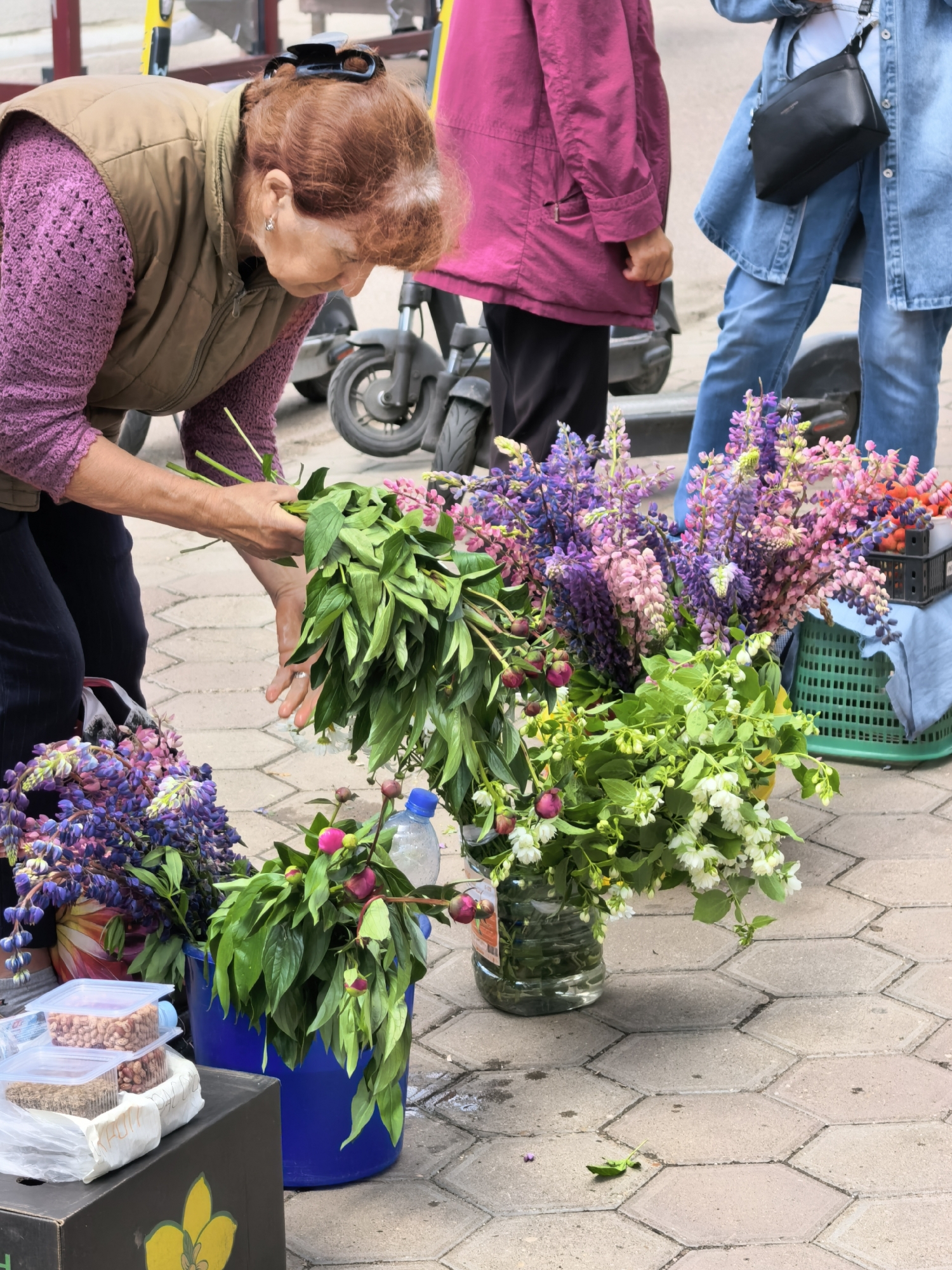 An elderly woman with red hair, wearing a brown vest over a purple knitted sweater and dark pants, is seen from the chest up, tending to a large bouquet of greenery in her hands. Her head is bowed, focused on her task. Behind her, partially visible, is another woman in a vibrant pink jacket and a person wearing a denim jacket and jeans, holding a black purse. The scene appears to be an outdoor market or street stall, with various bouquets of flowers, including purple lupine and pink peonies, displayed in buckets and baskets. Several scooters are visible in the background, suggesting an urban setting. The ground is paved with interlocking hexagonal stones. The presence of these elements indicates this is likely Sterlitamak, Russia, given the context provided.