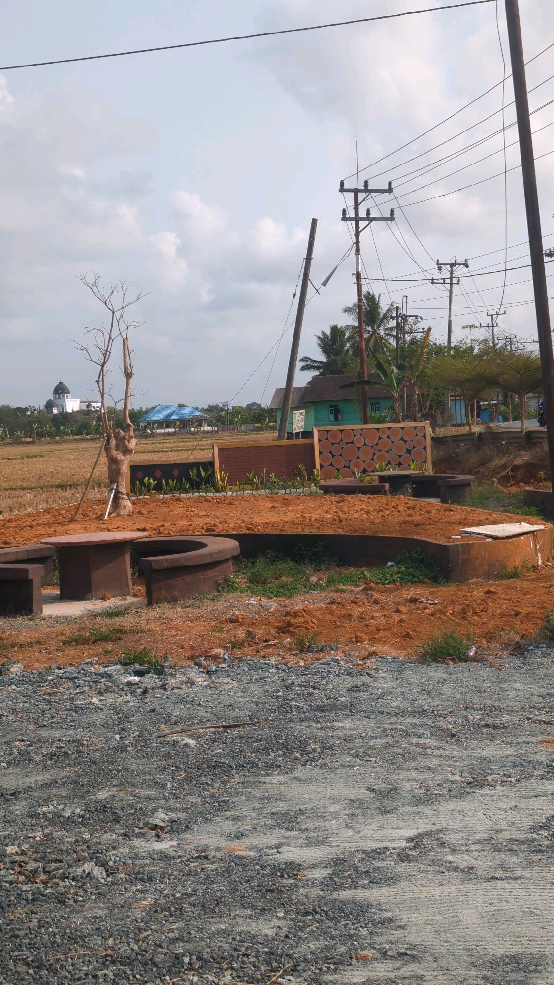 The image depicts an outdoor scene in Sepunggur, Indonesia, under an overcast sky. The foreground is dominated by a gravelly path leading to a construction or landscaping area. This area features reddish-brown soil, concrete seating structures, and planters filled with greenery. A bare, gnarled tree stands prominently in the middle ground, beside a circular concrete planter. Behind this, a decorative wall with a circular wooden pattern is visible, along with other brick and concrete elements.
In the background, several buildings can be seen, including a light green structure with a dark roof, and a blue-roofed building. A white building with a dark dome is visible in the far distance on the left. Power lines and utility poles crisscross the upper portion of the image. The landscape beyond the immediate area appears to be agricultural fields, possibly rice paddies, with palm trees scattered throughout. No people are visible in the frame.