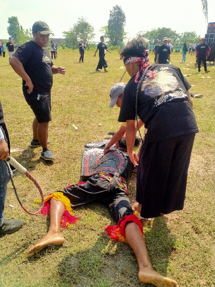This image depicts an outdoor scene in Celep, Indonesia, under daylight conditions. A person lies supine on a grassy field, barefoot, dressed in dark pants with prominent red and yellow fringed decorations at the ankles and a multi-colored, patterned chest covering. Two individuals are positioned over the person on the ground: one, wearing a grey baseball cap and dark t-shirt, leans forward towards the chest area; the other, wearing a dark t-shirt with a graphic on the back, dark wide-legged pants, and a red and white patterned bandana around their head, also leans over the prone individual. A dark braided rope or whip is visible near the second individual. To the left, a man in a dark t-shirt, dark shorts, and a baseball cap stands observing, with his hands on his hips. In the far left foreground, the lower body of another individual is visible, holding a whip with a braided handle. The background features an expansive grassy area with several individuals scattered throughout, mostly dressed in dark casual clothing. Trees line the horizon, and a large black sound system is visible on the far right. The ground consists of green grass with exposed patches of dirt.