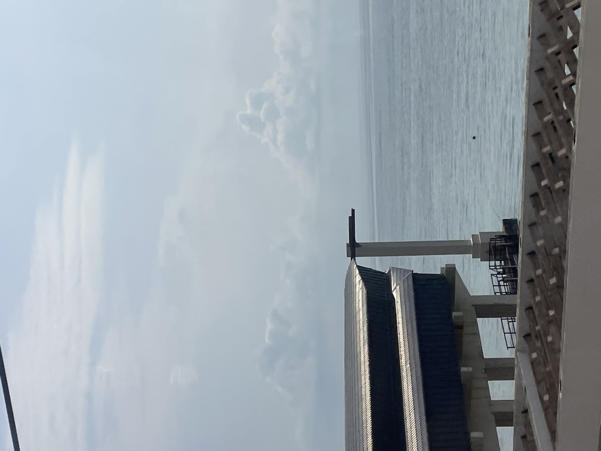 This is a vertical, eye-level, full-shot, outdoor photograph of a pier extending into the sea. The sky is overcast with fluffy white clouds, and the sea is calm and blue. In the foreground, there is a grey concrete barrier with an intricate pattern. The middle ground features a large, dark blue building with a metallic roof, supported by concrete pillars over the water. A white pillar with a horizontal beam extends from the building over the sea, suggesting a diving board or similar structure. A small red buoy floats in the water. The background shows the expanse of the sea meeting the hazy sky. This scene is located at the Olympic Park in Sochi, Russia, specifically the Sochi Autodrom area, which features a pier that extends into the Black Sea. There are no people visible in the image.