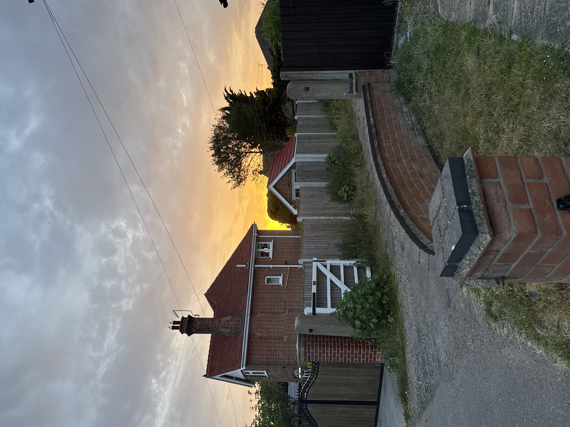 The photograph shows a residential street scene at dusk or dawn, likely in the Great Yarmouth area of the United Kingdom. A two-story brick house with a red tile roof occupies the left side of the frame. The house has a brick chimney, a few small windows, and a white-painted gate. A wooden fence with a small red roofed shed behind it and green foliage runs alongside the house. To the right of the fence, there's a brick and concrete pathway leading towards the foreground. A brick and concrete pillar stands on the right edge. Power lines extend across the upper portion of the frame. The sky is a mix of grey clouds and a warm, golden hue from the rising or setting sun.