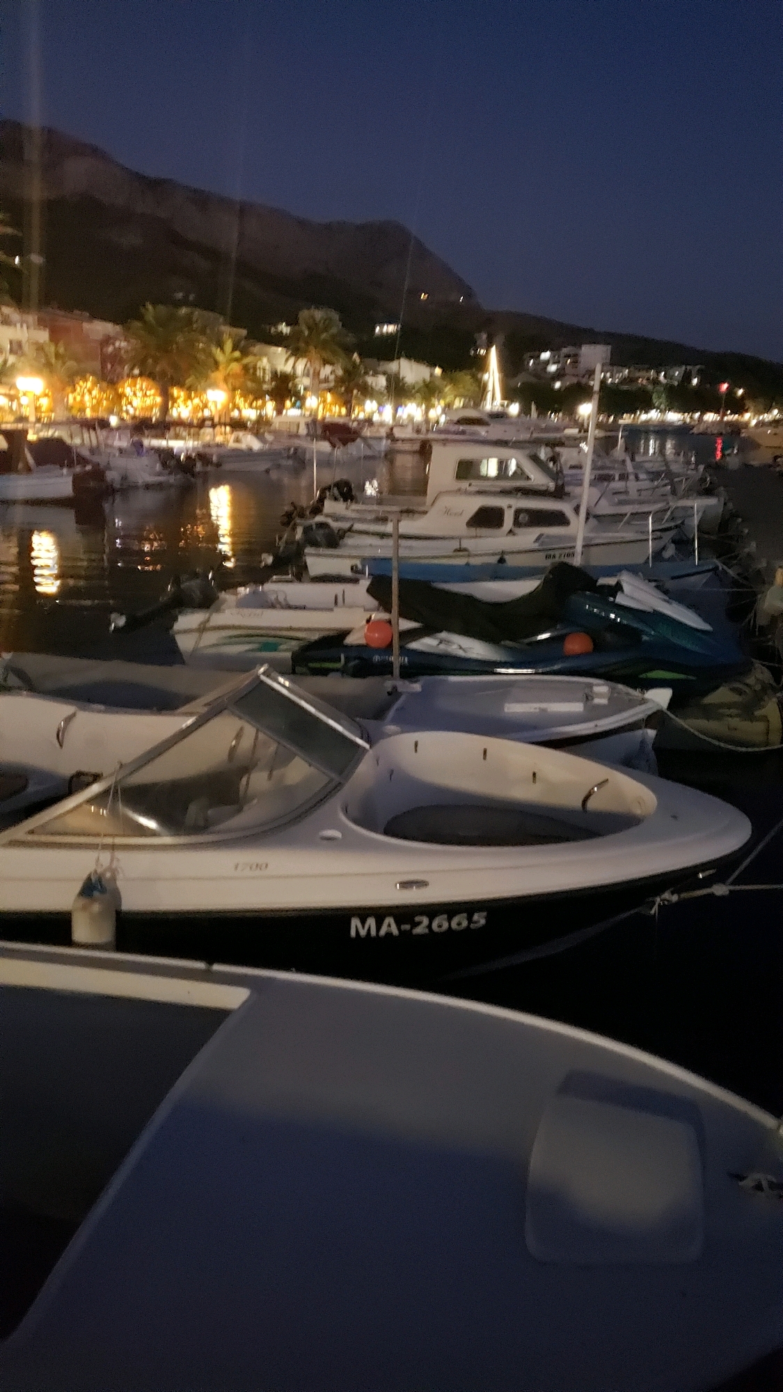 This is a nighttime, vertical, medium shot of a harbor in Općina Tučepi, Croatia. In the foreground, two boats are docked. The boat closest to the viewer is a white speed boat with a dark hull. The registration number "MA-2665" is visible on its hull. Behind this boat, another speed boat is docked, partially obscured by the first. Further back, multiple other boats, including a jet ski, are visible. In the background, a town with illuminated buildings and palm trees lines the coast, with a mountain range behind it. The sky is dark blue and clear.