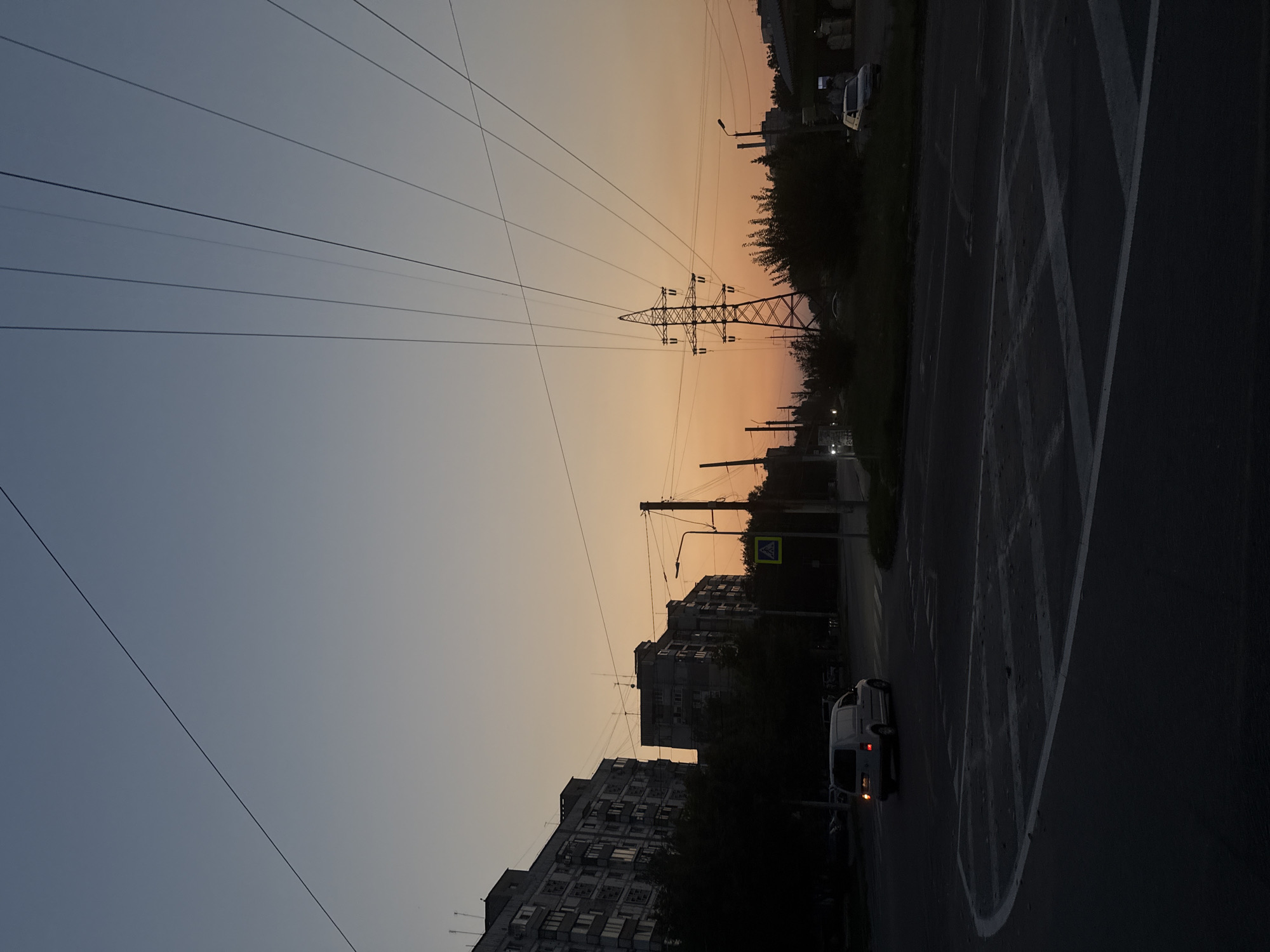 The image depicts a street scene at dusk in Zaporizhzhia, Ukraine. A light-colored van is turning at an intersection, its brake lights illuminated. The sky exhibits a gradient of colors, transitioning from a pale blue near the top to a warm orange and pink near the horizon. Multiple power lines stretch across the sky, converging towards a large electrical pylon on the right side of the frame. Tall apartment buildings, characteristic of Soviet-era architecture, line the left side of the street. A pedestrian crossing sign is visible on a pole to the right of the intersection. The road surface is dark and appears to be asphalt, with white markings indicating lanes and the crosswalk. Trees and bushes are sparsely scattered along the roadside. The overall impression is a quiet, urban setting during the evening twilight.