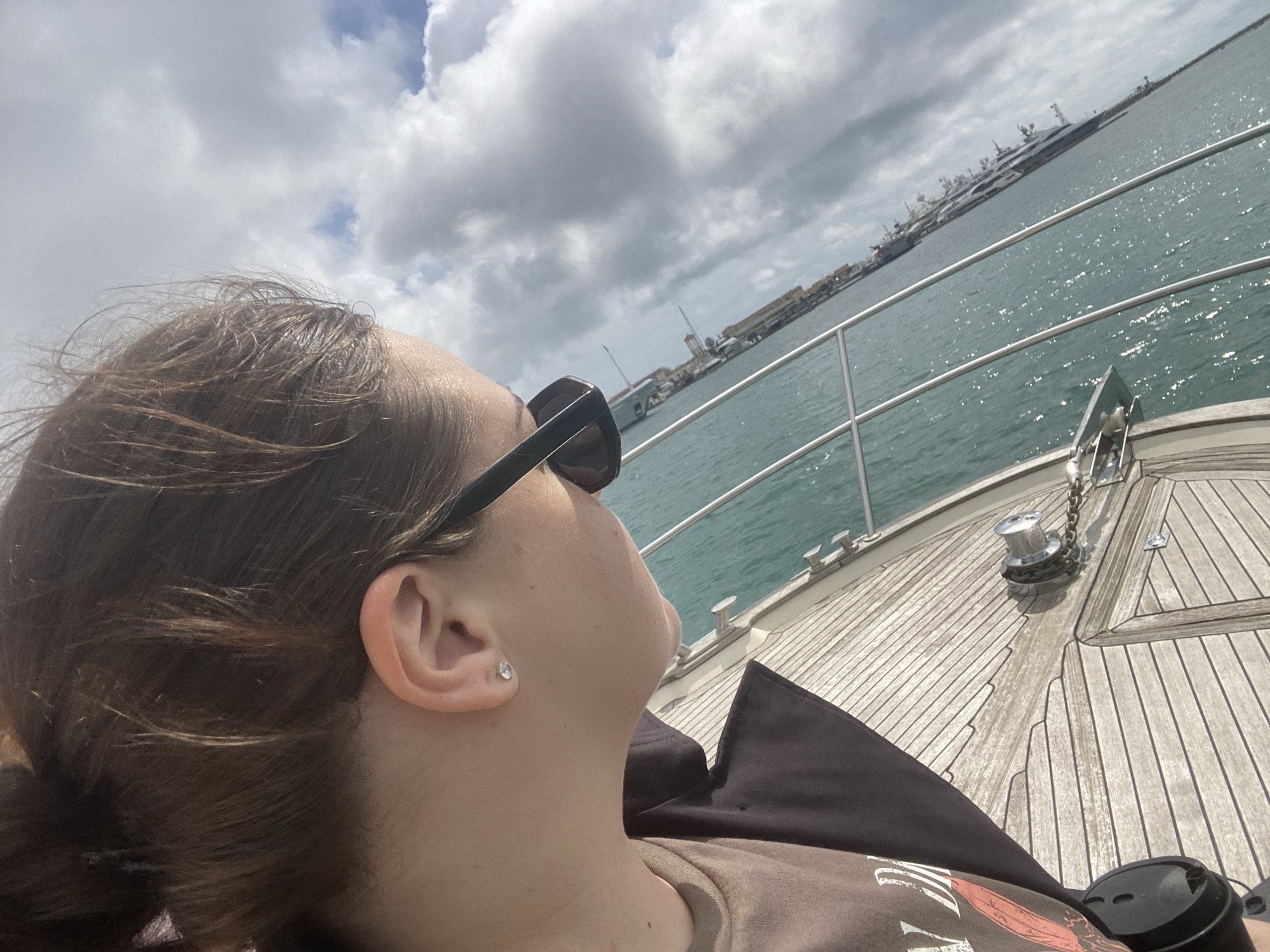 A woman with sunglasses is lying on a boat deck, looking up at the sky. The deck is made of wooden planks. A railing runs along the edge of the boat, and beyond it is a body of water. In the background, there are buildings and other boats. Given the presence of yachts, the scene may be occurring near the Sochi Grand Marina in Sochi, Russia.