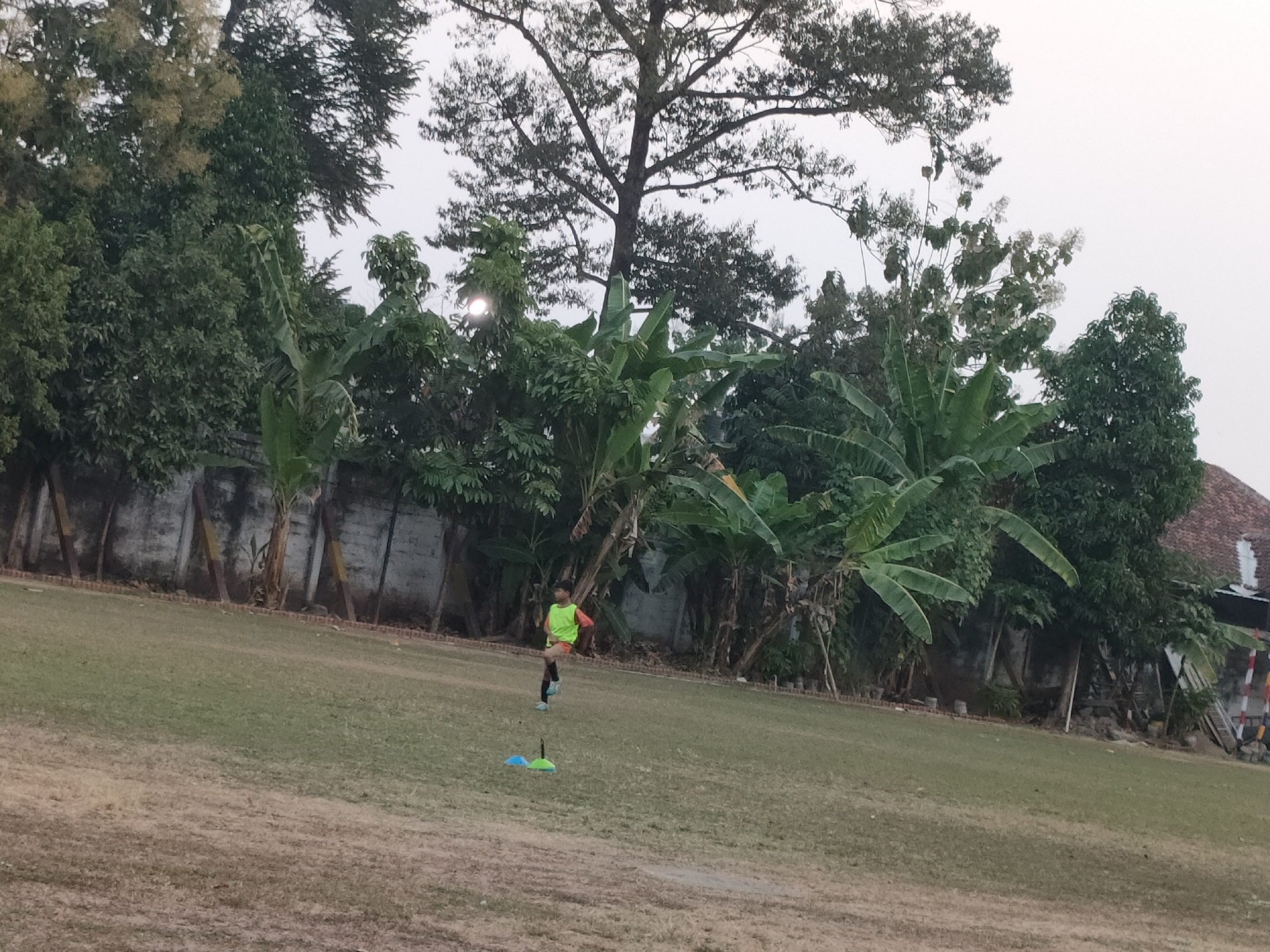 In Banguntapan, Indonesia, on a grassy field, a young boy in a bright yellow vest and dark shorts runs past two colored cones, one blue and one green. The field is surrounded by a line of small potted plants and a low brick wall, behind which are trees and a building with a tiled roof. The sky is overcast.