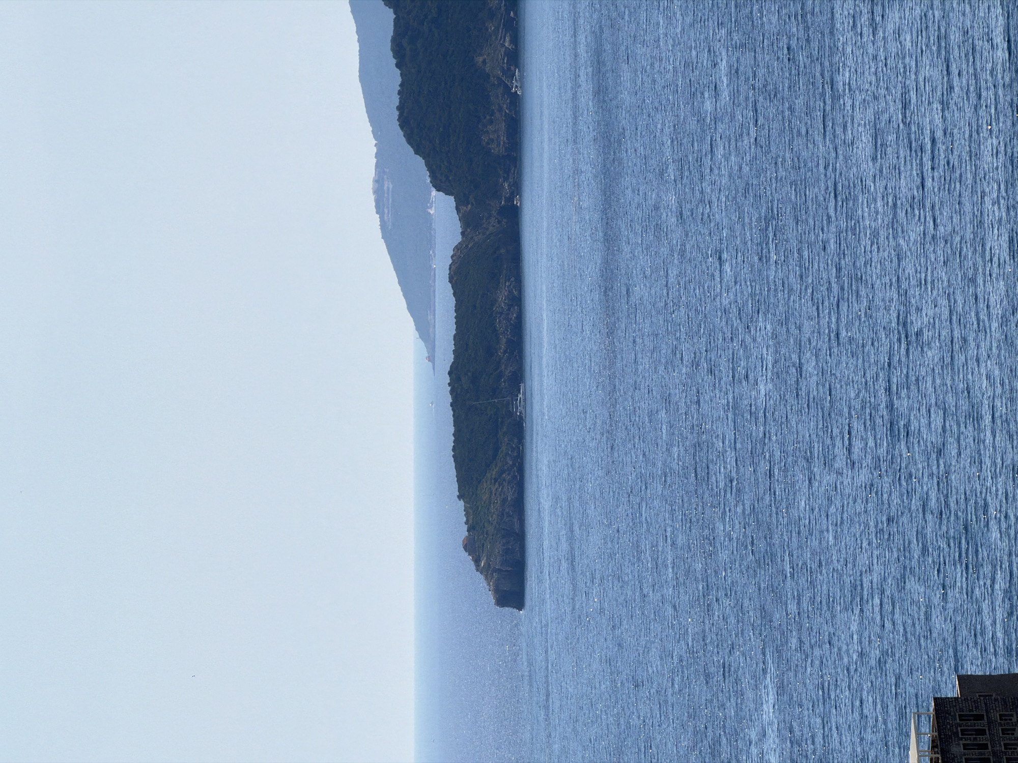 The image depicts a scenic coastal view of the Adriatic Sea near Sveti Stefan, Montenegro. The foreground is dominated by the deep blue, rippling surface of the sea. In the mid-ground, a rugged, tree-covered peninsula juts out into the water, with a small building visible on its rocky summit. Further in the distance, hazy green hills and mountains form the backdrop, suggesting a mountainous coastline. Two white yachts are visible in the water near the peninsula. On the far left of the image, a section of a stone building with an awning can be seen, suggesting structures built along the coast. The sky is a pale, clear blue, indicating a bright, sunny day.