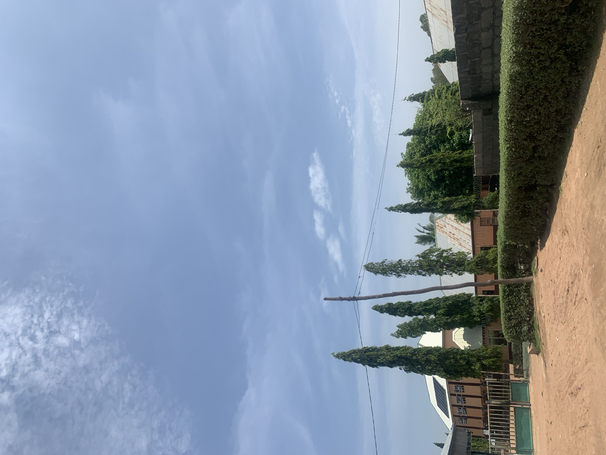 The image depicts a view of buildings and vegetation under a partly cloudy sky. In the foreground, a dirt road is visible, bordered by a neatly trimmed green hedge. Behind the hedge, a dark grey brick wall runs horizontally across the frame. Beyond the wall, several tall, slender evergreen trees stand out against the sky, interspersed with other trees and foliage.
Further back, a light-colored building with a corrugated metal roof is visible. Some sections of the roof appear aged and worn. On the roof of one of the buildings, solar panels are installed. The buildings have windows and architectural features consistent with residential or institutional structures. A green metal gate is partially visible on the left side of the image, suggesting an entrance. A wooden pole with electrical wires strung between it and other unseen poles is also present.
The sky is a bright blue with scattered white clouds, indicating daytime. The overall scene suggests a tranquil environment, possibly a residential area, school, or compound in Jauro Yinu, Nigeria. No people are visible in the image.