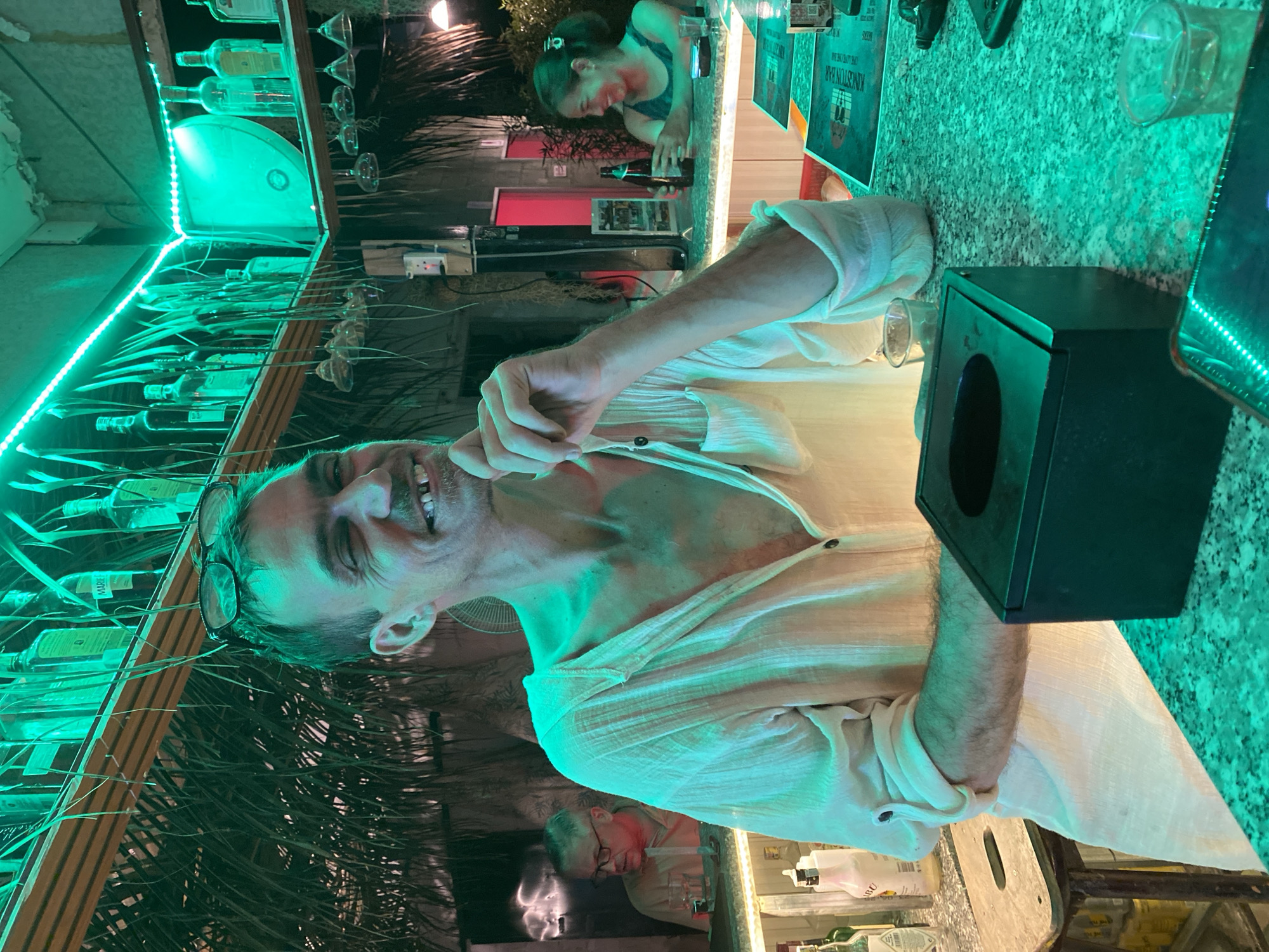 The image depicts an indoor bar scene in Mũi Né, Vietnam, characterized by dominant green and teal lighting.
In the foreground, an adult male with short dark hair and glasses pushed up on his forehead is seated at a bar counter. He is smiling widely, with his eyes partially closed and a noticeable gap between his two upper front teeth. He wears a loose-fitting, light-colored (off-white or cream) long-sleeved shirt, unbuttoned several buttons down the chest, revealing chest hair. His sleeves are rolled up. His right hand is raised to his chin, with fingers resting near his mouth, while his left arm rests on the counter. In front of him on the bar counter is a black tissue box holder and a small, clear shot glass. A reflective surface, possibly a smartphone, is visible at the bottom edge of the frame.
The bar counter surface is speckled green and white, appearing to be granite or a similar stone. Behind the man, various liquor bottles are arranged on a lower shelf, including one labeled "MALIBU." A rustic wooden stool is partially visible to the left.
In the mid-ground, a second adult male wearing glasses is seated behind the main subject, also at a bar. He wears a light-colored shirt and appears to be looking to his left. Further back and to the right, a woman with dark hair pulled back is seated at another section of the bar. She is smiling and holding a dark glass bottle, possibly a beer, with both hands.
The bar's overhead structure features shelves made of wood, from which numerous stemmed glasses hang upside down. The shelves also hold various clear and dark glass liquor bottles, with one visible bottle bearing the label "MARIE." Green LED strip lighting illuminates the area above and behind the shelves, casting a strong green glow throughout the scene. Decorative thatch-like material and dried grass elements hang from the ceiling structure, contributing to a tropical aesthetic. A white electrical outlet is mounted on a column or wall in the mid-ground. A small, circular wall-mounted fan is visible to the right of the main subject. In the far background, a doorway emits a faint red glow, and some foliage is visible behind the woman. A menu or sign on the bar counter to the right clearly displays the text "KONSTANTIN BAR."