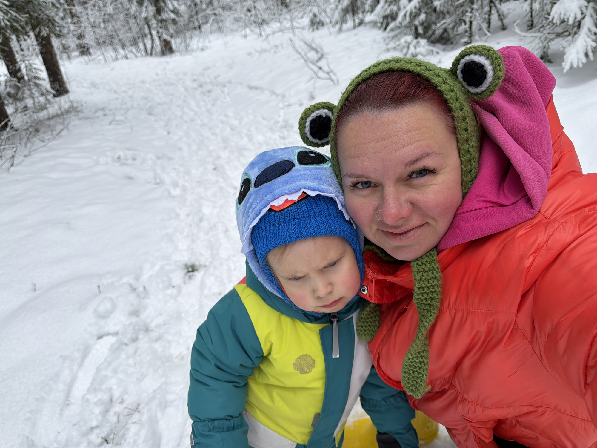 The image depicts a woman and a young child in a snowy outdoor environment. The woman is wearing an orange puffer jacket with a pink hoodie underneath, and a green crocheted hat with large frog-like eyes. The child is wearing a blue and yellow snowsuit with a blue hat featuring a shark-like design. They appear to be standing on a snow-covered path in a wooded area, with trees visible in the background. The location is Chaikovsky, Russia.