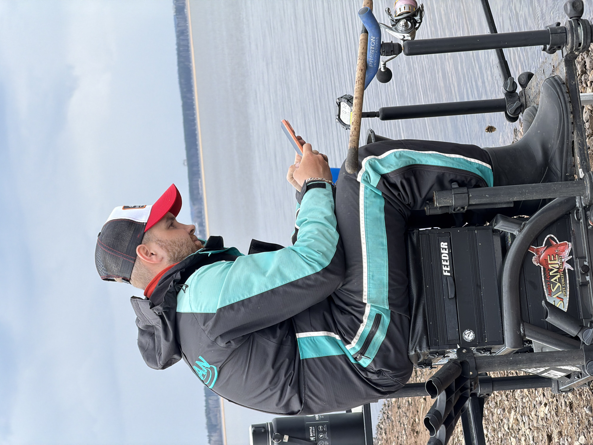 A man wearing a red, white, and grey baseball cap, a grey and turquoise jacket, and black boots is seated in a fishing chair by a body of water. He is looking at and holding a cell phone. Fishing rods are set up in front of him. The fishing chair is black, with the words "FEEDER" and "SASAME" visible on it. The location is Chaikovsky, Russia, indicated by the jacket having a logo with the Russian letter "Ч". The shoreline is visible in the foreground.
