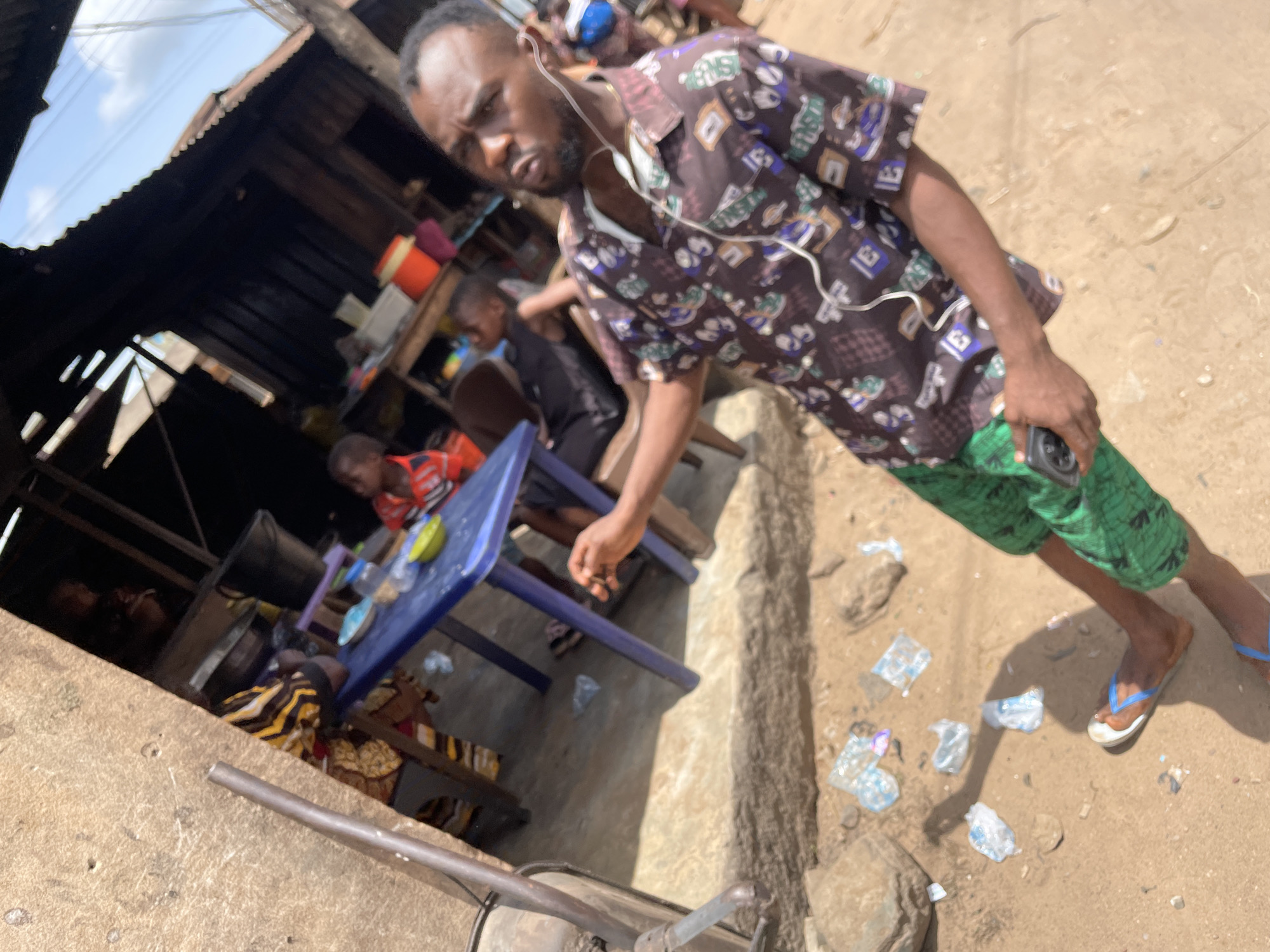 In Onitsha, Nigeria, an outdoor scene shows a man holding a phone and wearing a patterned shirt, green shorts, and flip-flops. He has earphones connected. To his left, people are seated at a blue table. A small cart is visible in the foreground. The environment appears to be a semi-urban or market area. The ground has scattered debris. A structure with a corrugated roof provides shade to the left.
