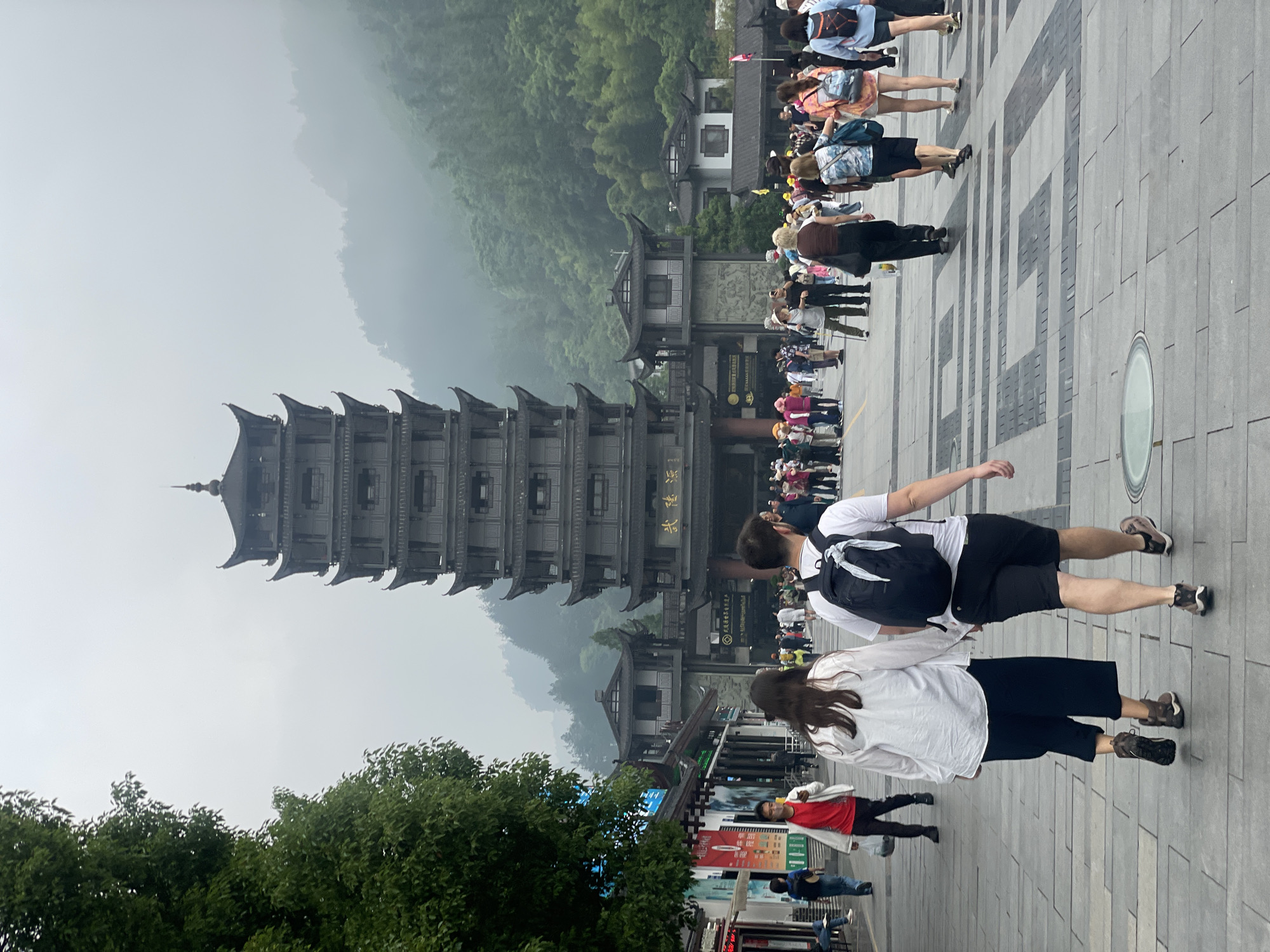 The image shows a tall pagoda-like structure in Wulingyuan, China, with many tourists in front of it. The structure is multi-tiered and of dark gray color. It stands behind a large entrance gate with a traditional Chinese roof design. Lush green mountains rise behind the structure and are covered in mist. A few people wearing casual clothes are walking on a paved plaza in the foreground, away from the pagoda and away from the rest of the people. There are green trees on the left side of the image. The plaza is made of square tiles arranged in a distinct pattern.