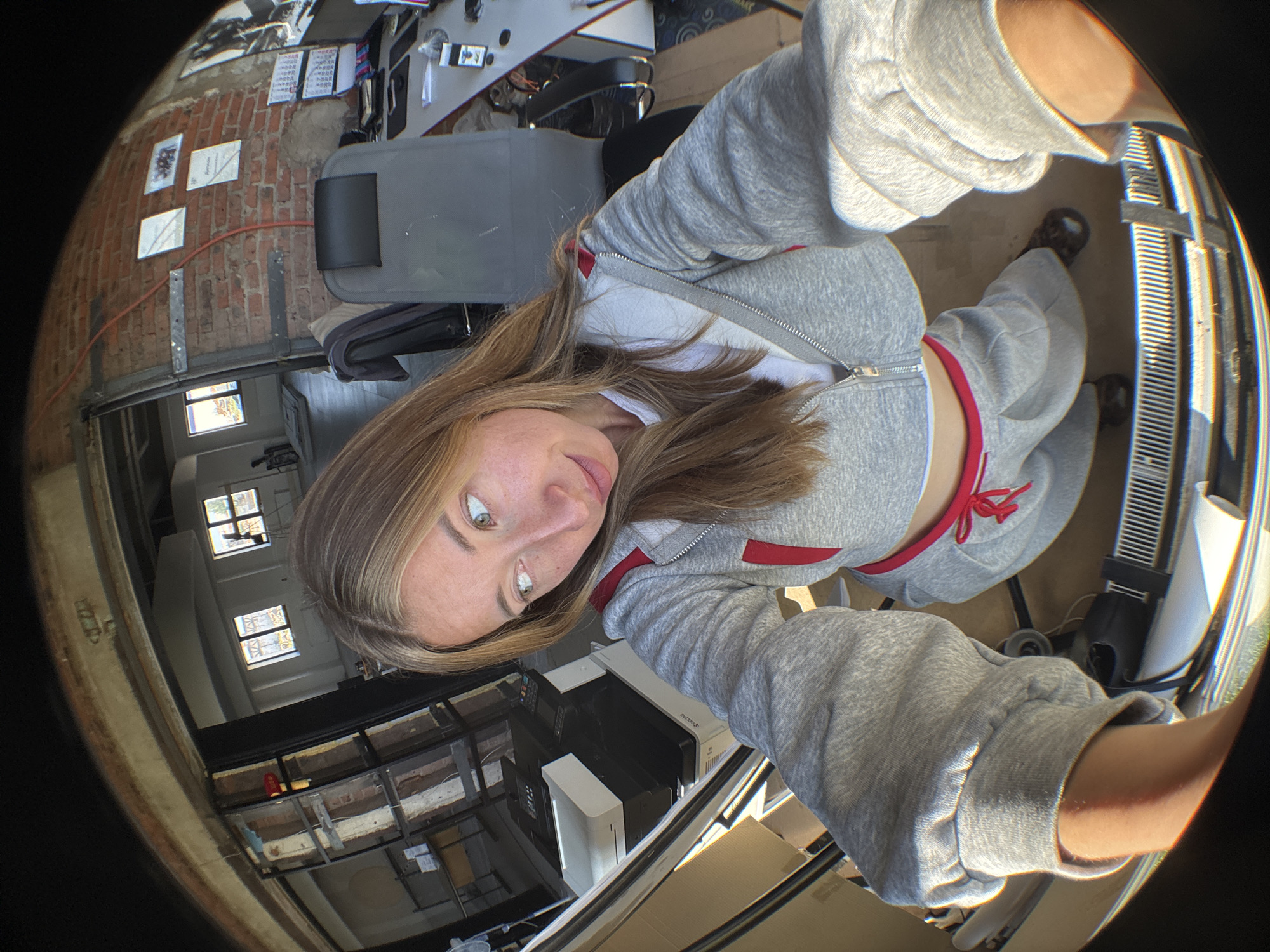 A young woman with blonde hair takes a selfie in an unfinished industrial space. She is wearing a grey zip-up hoodie, a white t-shirt, and grey sweatpants with red trim and a red drawstring. She is looking to her right with a slight smile. The background shows exposed brick walls, windows, an office chair, and a printer. The location is identified as Chaikovsky, Russia.