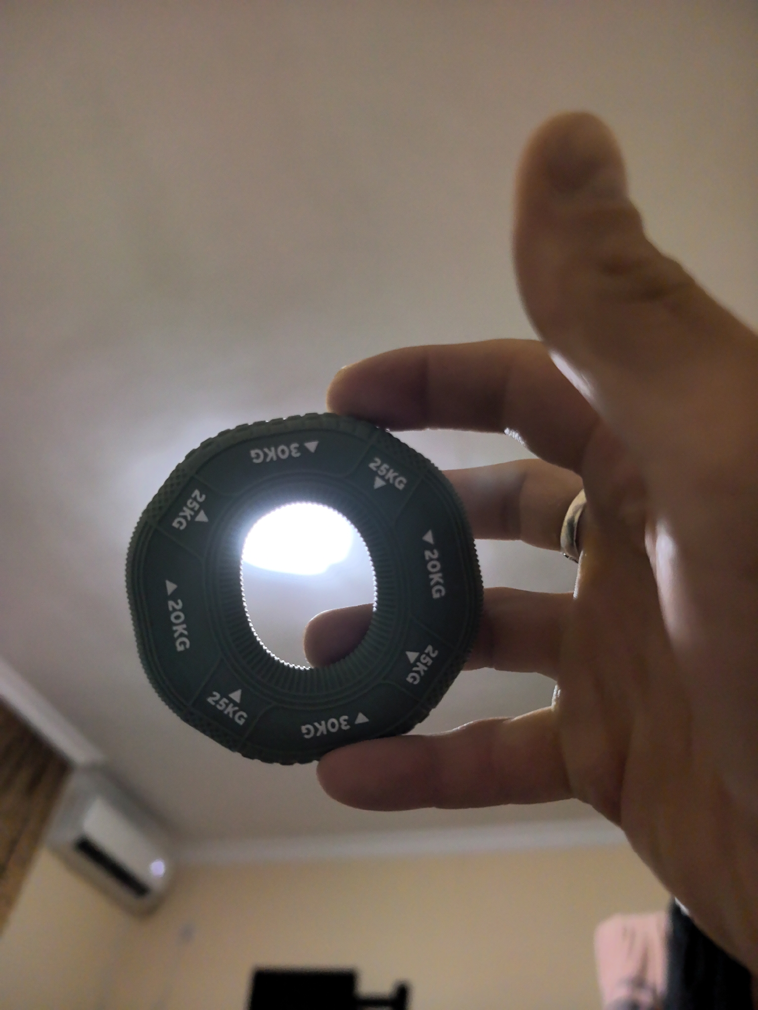 A hand is holding a green circular grip strengthener. The grip strengthener has markings around its circumference that read "20KG", "25KG", and "30KG". The background is blurred, but appears to be an indoor setting with a white ceiling and a wall with an air conditioning unit. The image does not provide any information to indicate the location in the city of Sochi, Russia.