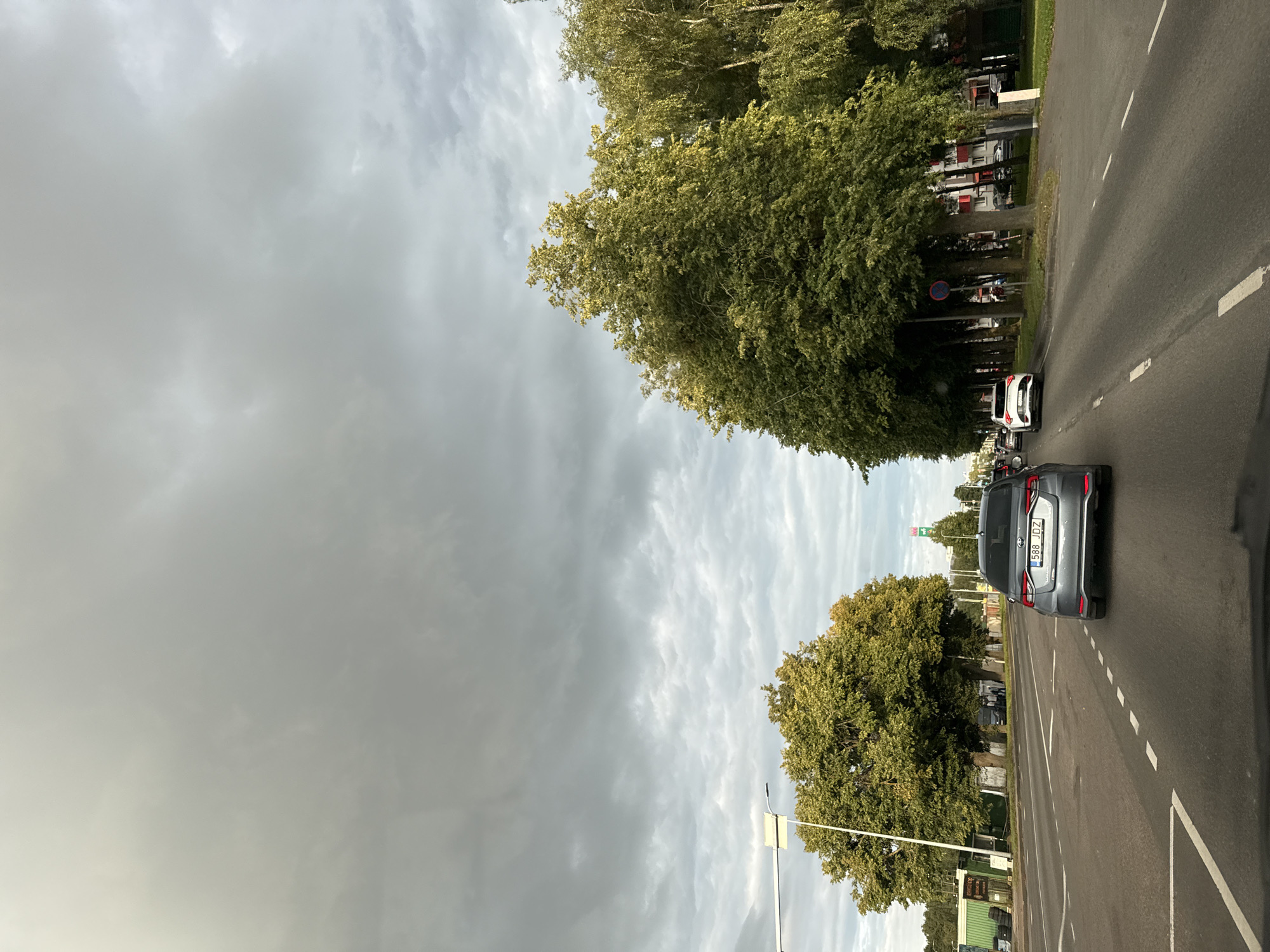 A grey car with license plate "588 JDZ" is driving on a multi-lane road in Tallinn, Estonia, under a cloudy sky. The car is in the right lane, with other cars visible ahead in both lanes. Tall trees line the right side of the road, and buildings are visible behind them. On the left side of the road, there are more trees and a building with green siding. A traffic light is visible on the left side of the road. The scene appears to be a city street with moderate traffic.