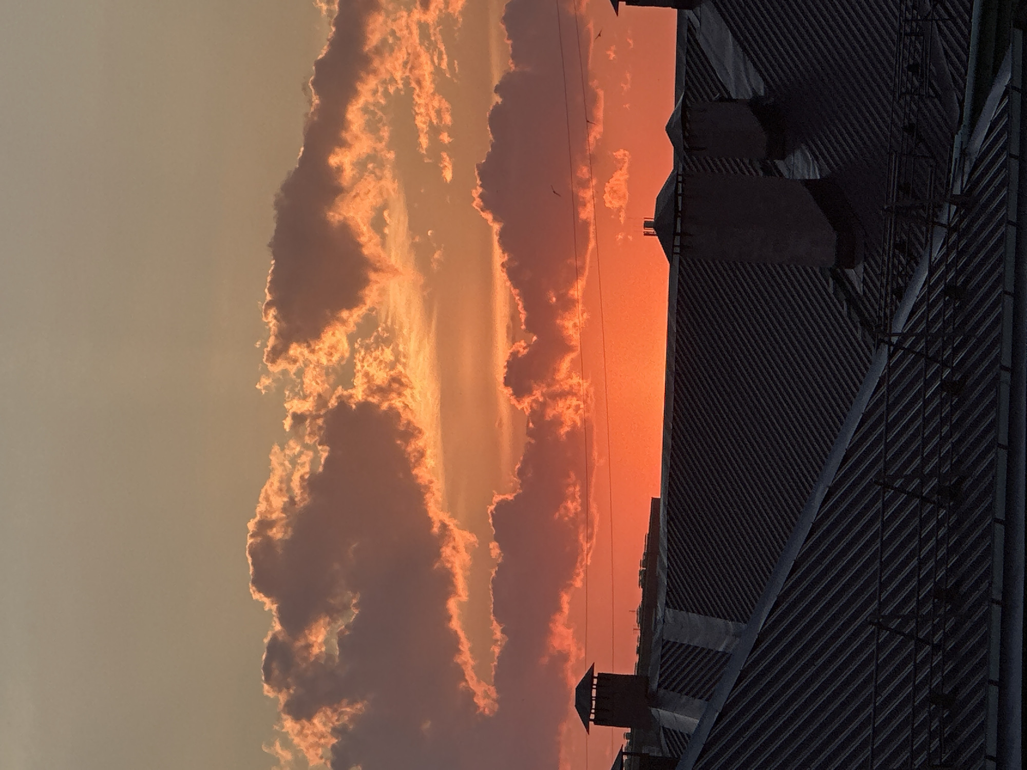 This is an outdoor, eye-level, medium shot of a sunset over a city. The sky is filled with clouds tinged with orange and red hues, indicating a vibrant sunset. Below the sky are rooftops of buildings, appearing dark and silhouetted against the bright sky. Several chimneys are visible on the rooftops. The image appears to be taken from a higher vantage point, looking down at the rooftops. There are no people visible in the image. The scene is serene and captures the beauty of a sunset in an urban environment. This image may have been taken in the city of Omsk, Russia, as requested.